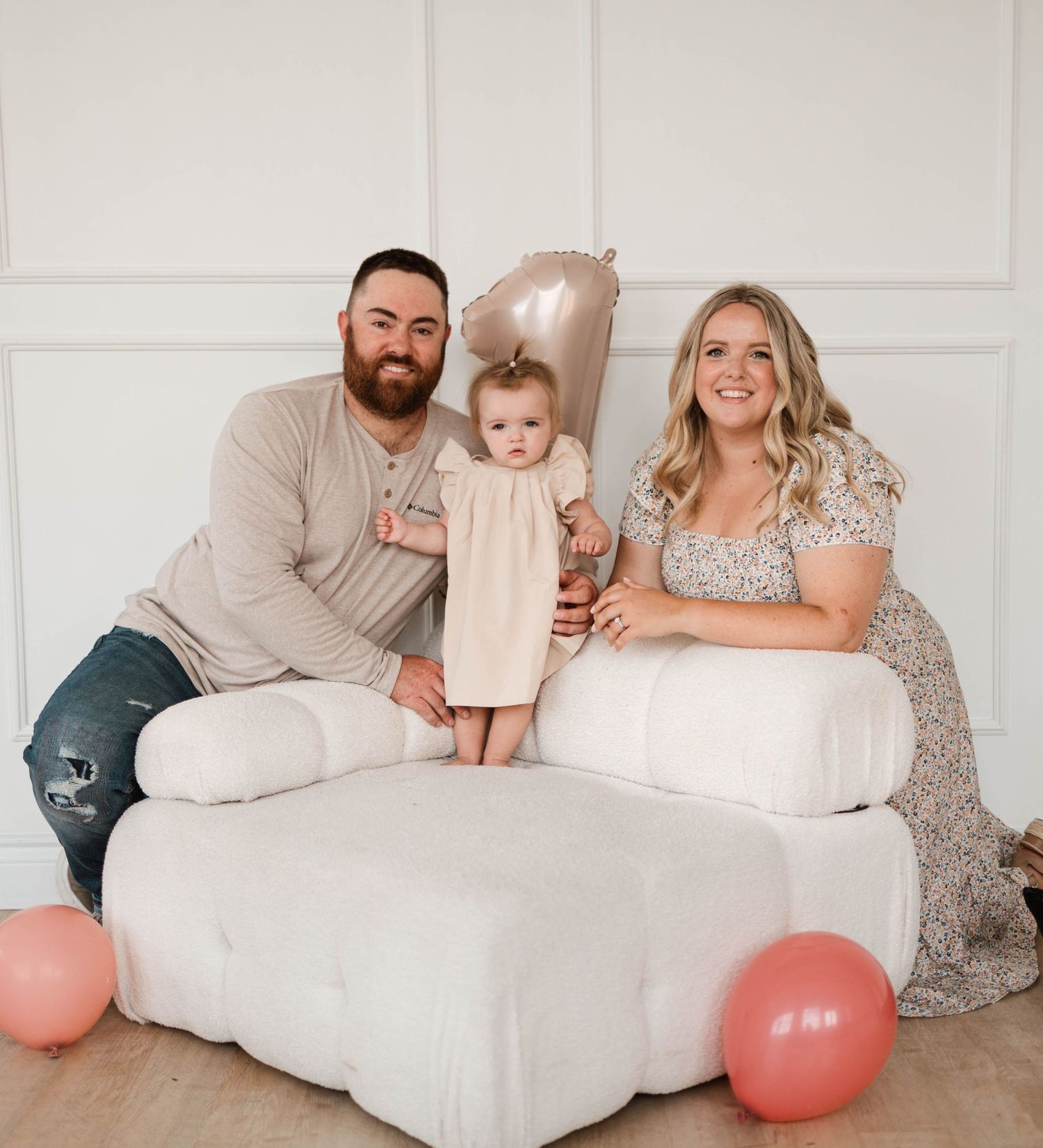 Family portrait with father, mother in floral dress, and baby girl with pink balloons indoors.