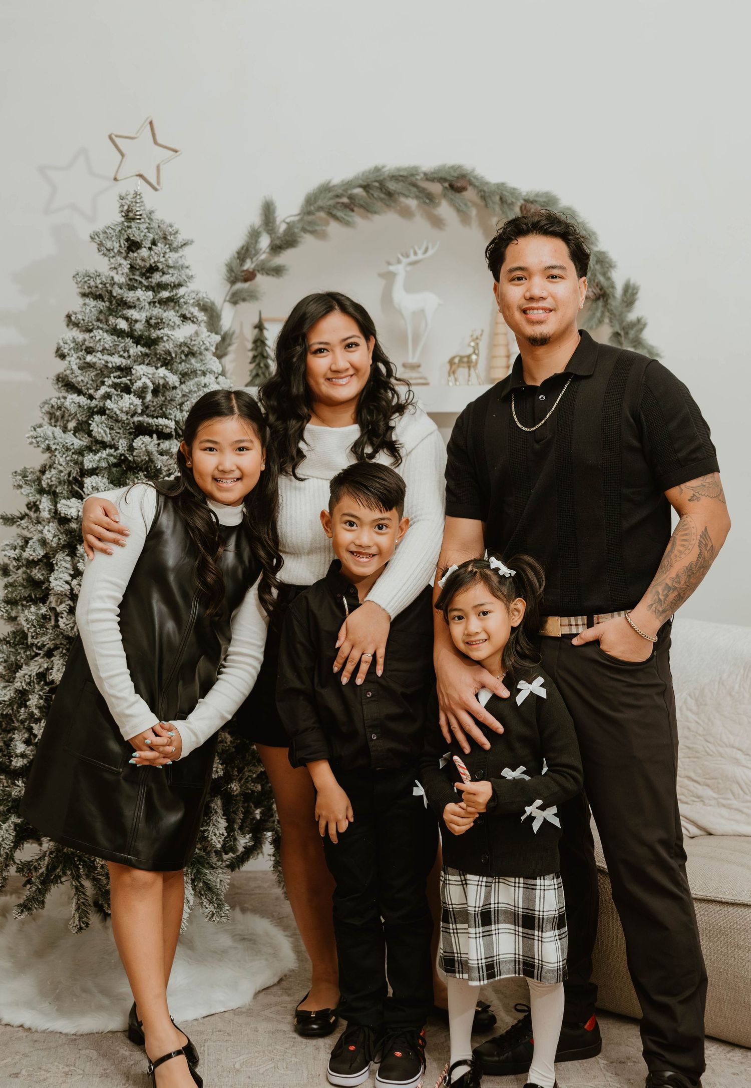 Family of five in coordinated black outfits posing by flocked Christmas tree indoors.