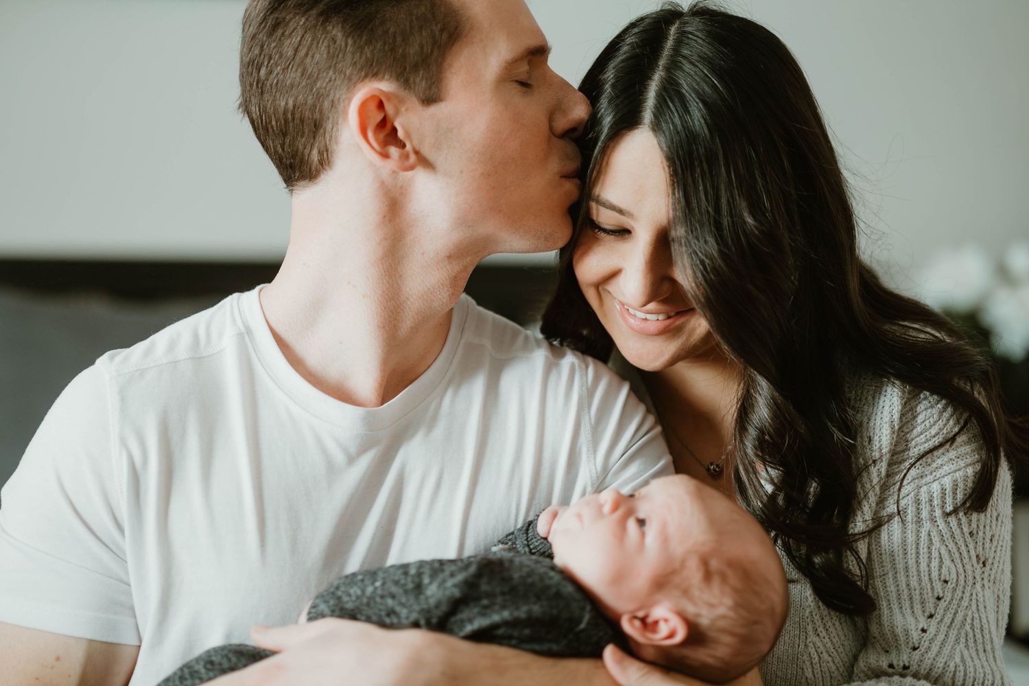 A dark-haired father kisses his smiling wife's head as she cradles their swaddled newborn baby.