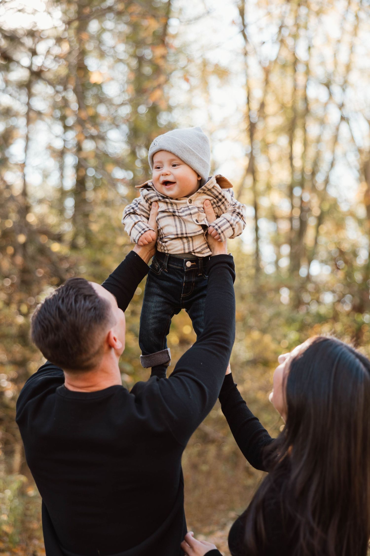 Parents lifting smiling baby in plaid jacket and grey hat up in the air in a sunny autumn forest.