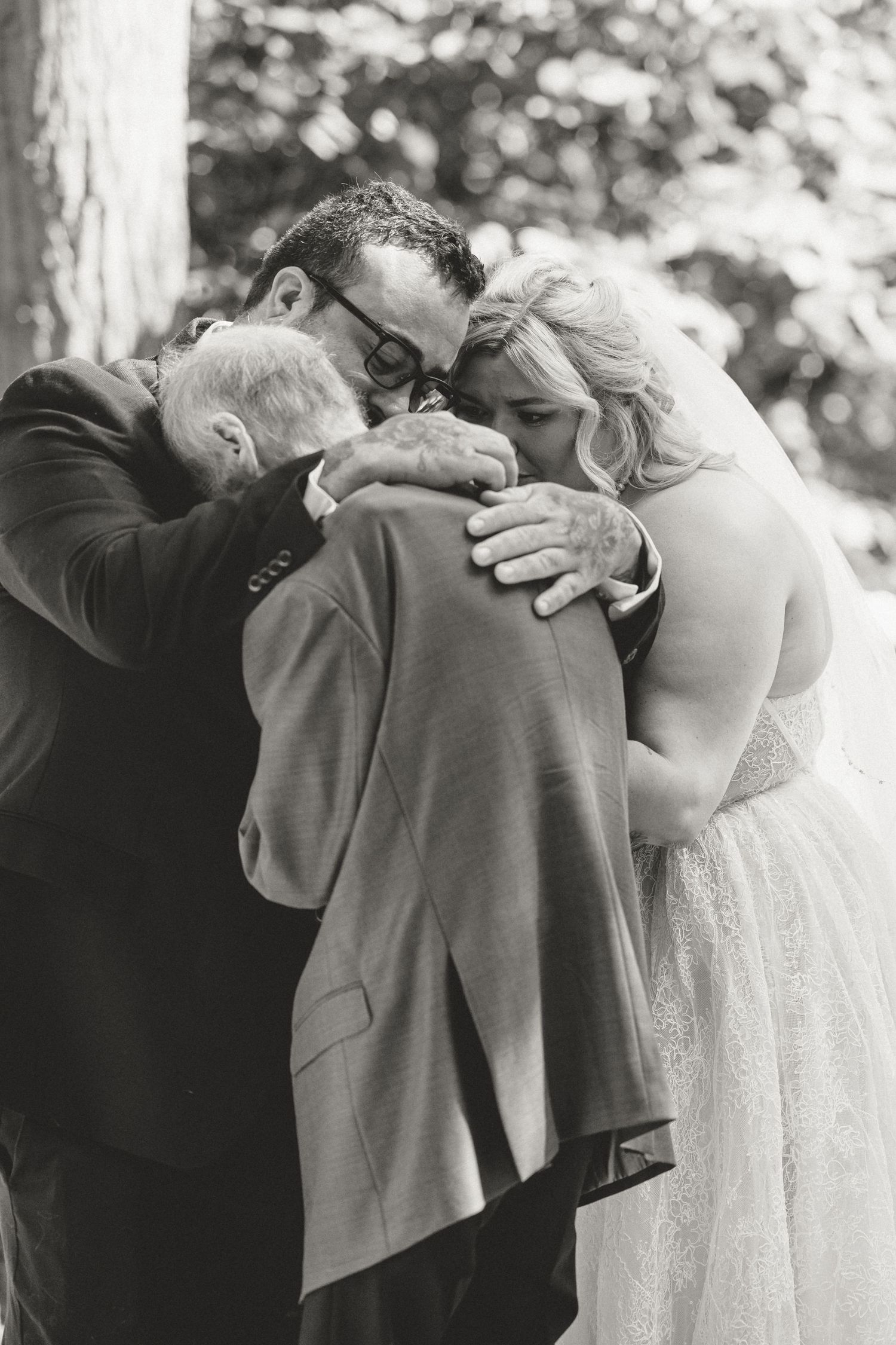 Black and white photo of bride, groom, and family sharing emotional group hug outdoors.