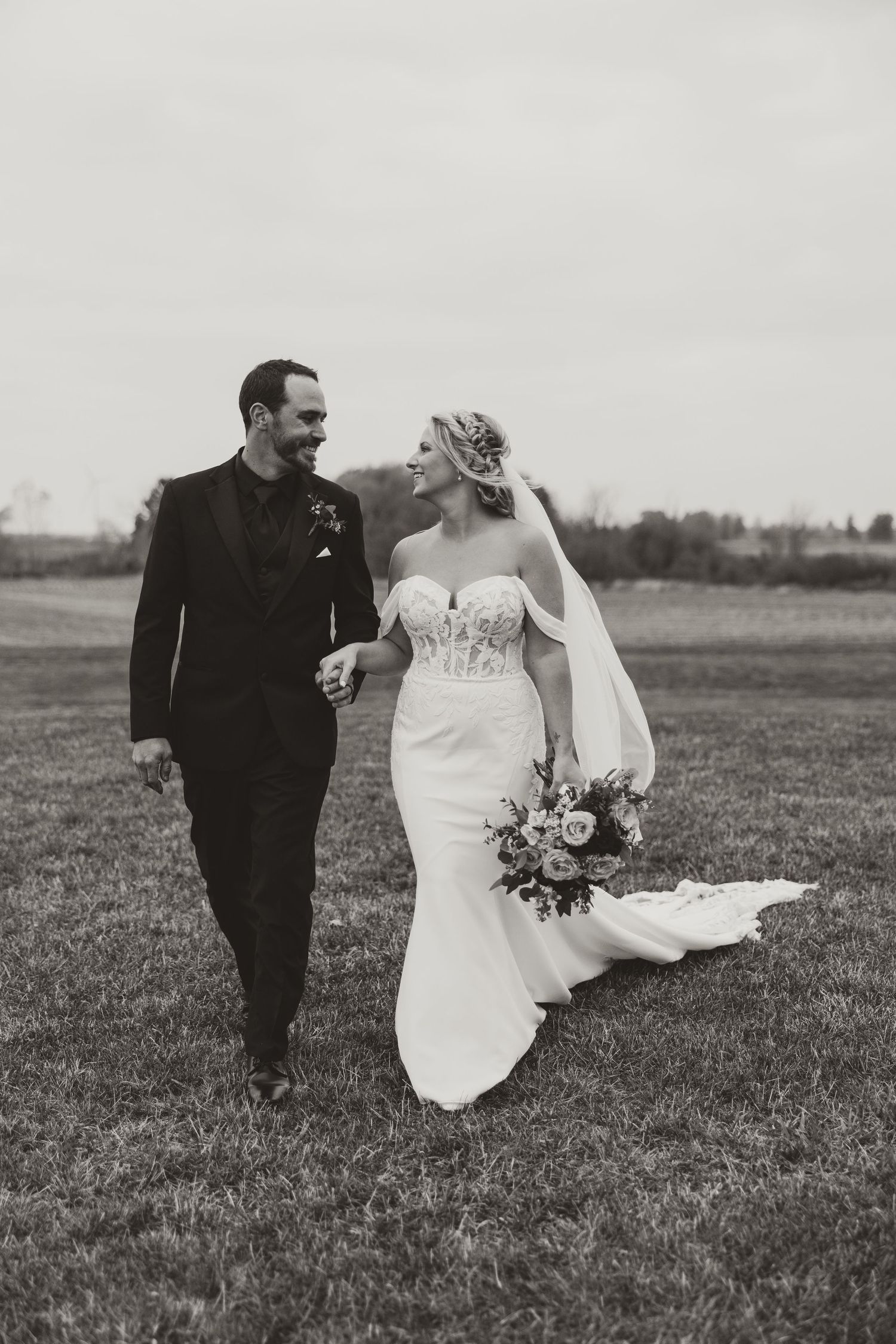 Black and white photo of smiling bride and groom walking hand in hand across a grassy field.