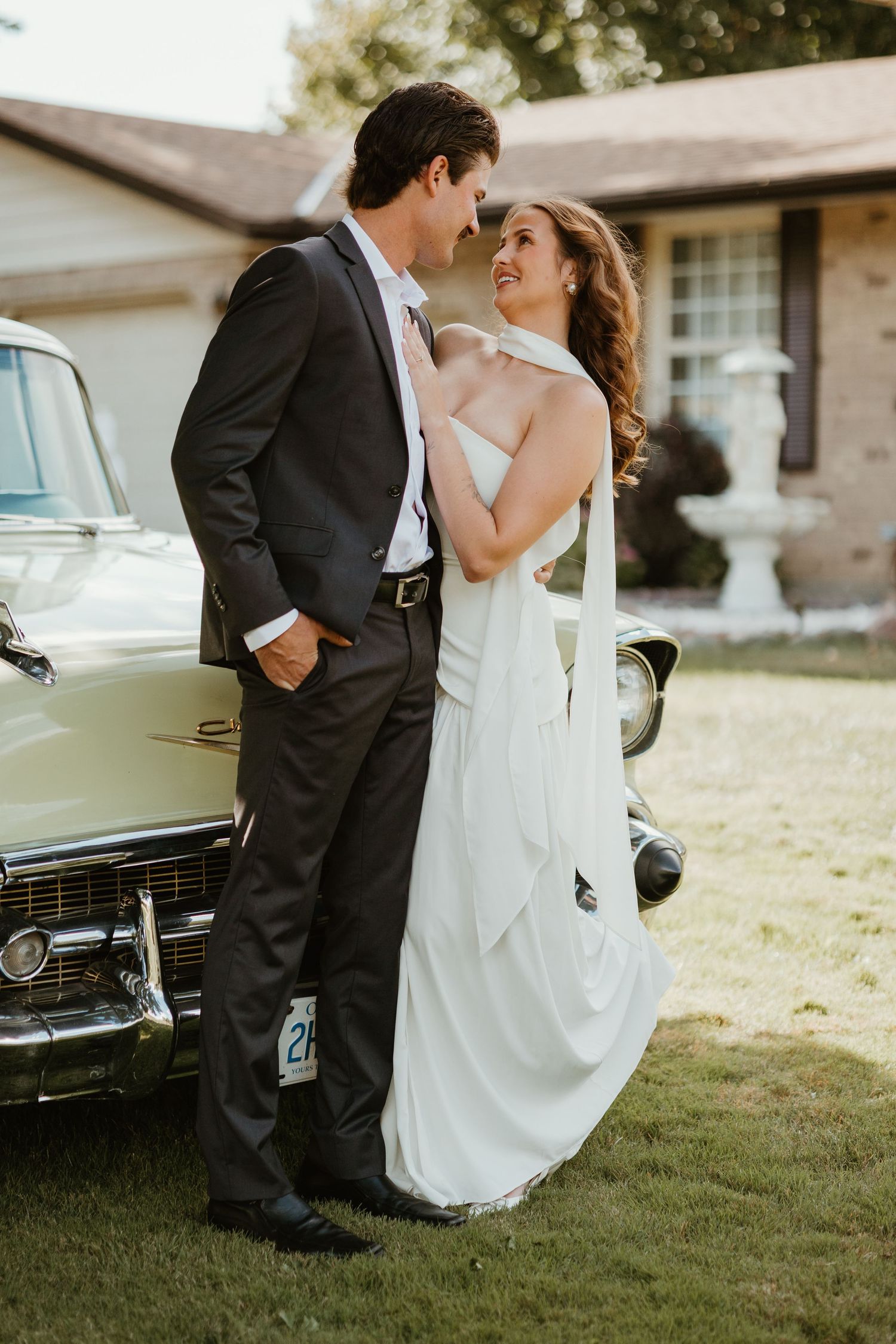 Bride and groom share a romantic moment leaning against a vintage yellow classic car on a sunny day.