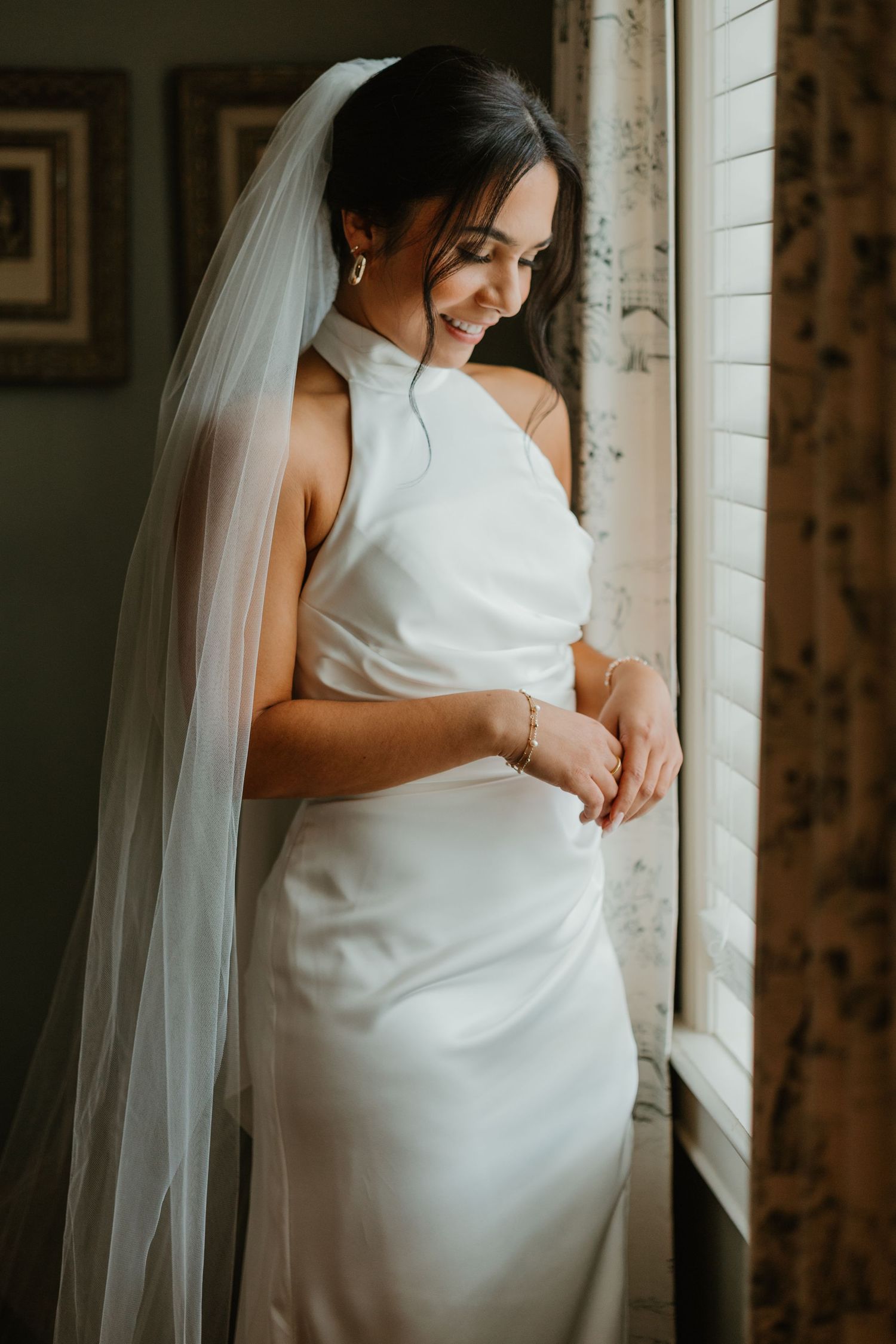 Elegant bride in sleek white halter gown and veil smiling by a sunlit window on her wedding day.