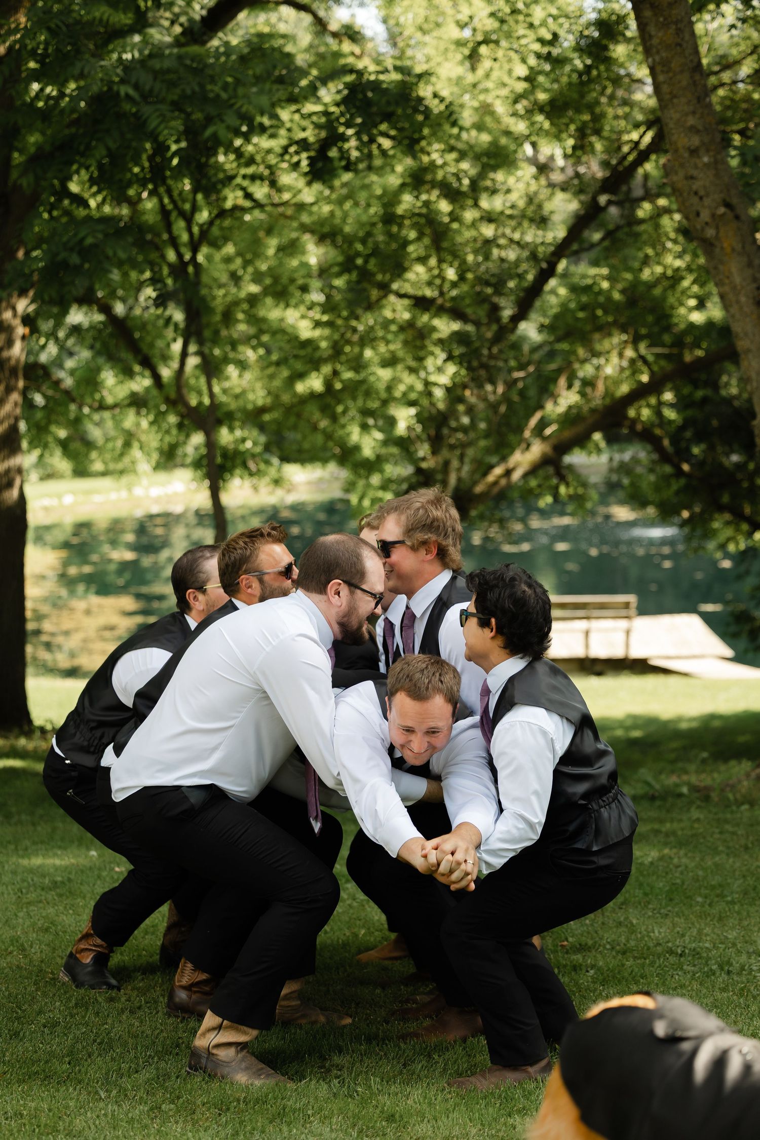 Groomsmen in black vests huddle together laughing on green lawn during outdoor wedding celebration.