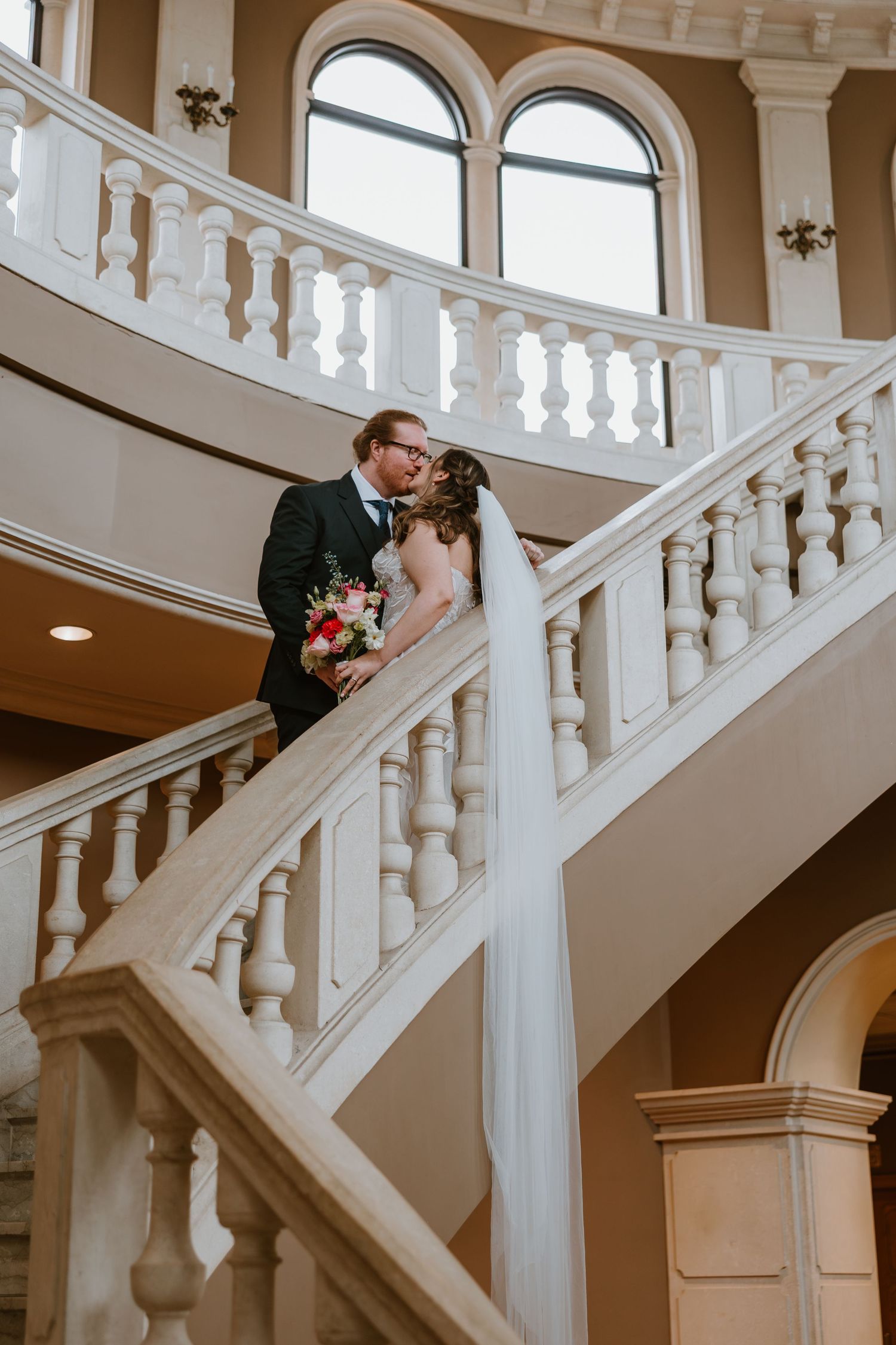 A bride and groom share a kiss on an elegant curved white staircase, bride holding a pink bouquet with flowing veil.