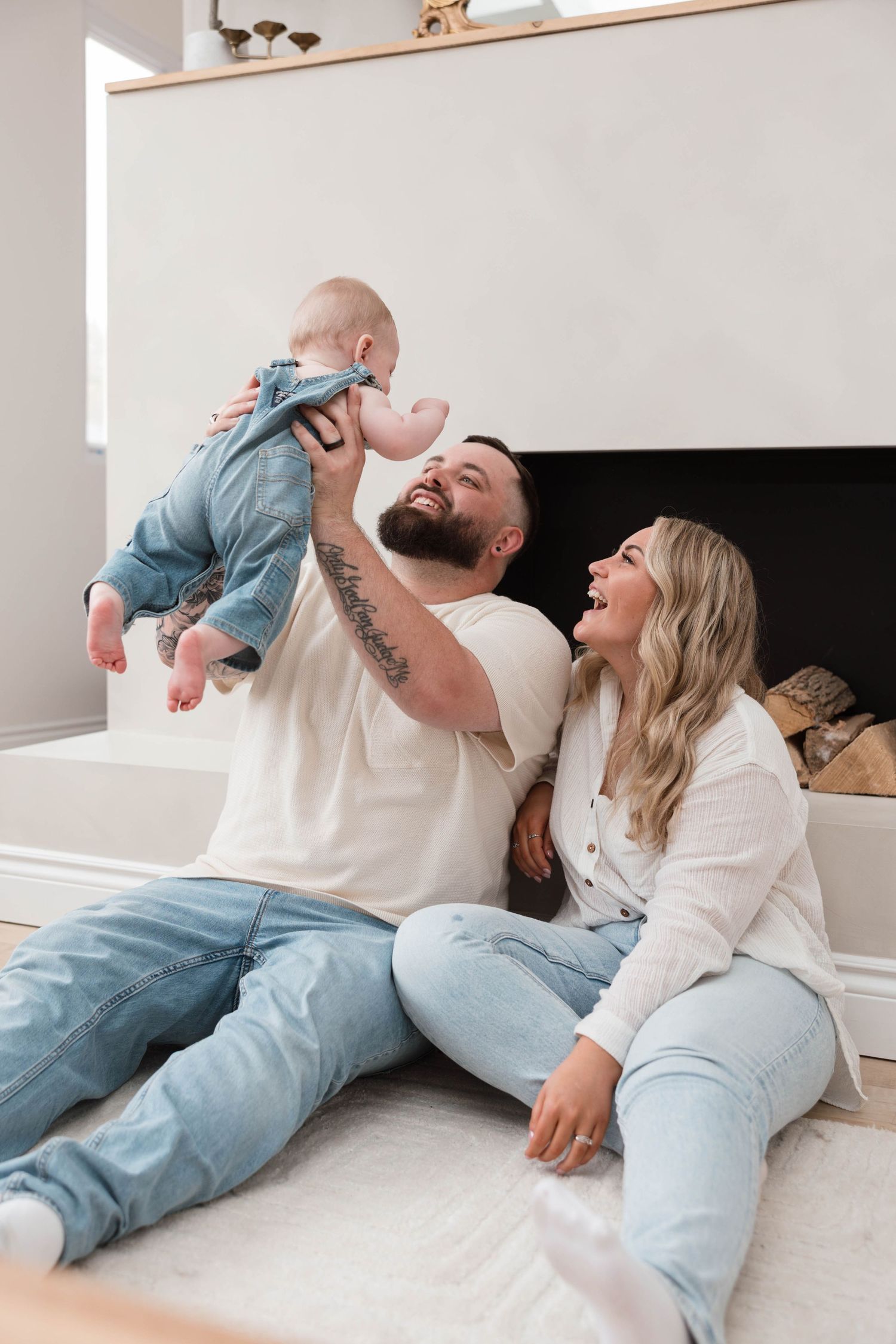 Happy family sitting on floor, father lifting baby in denim outfit near modern fireplace.