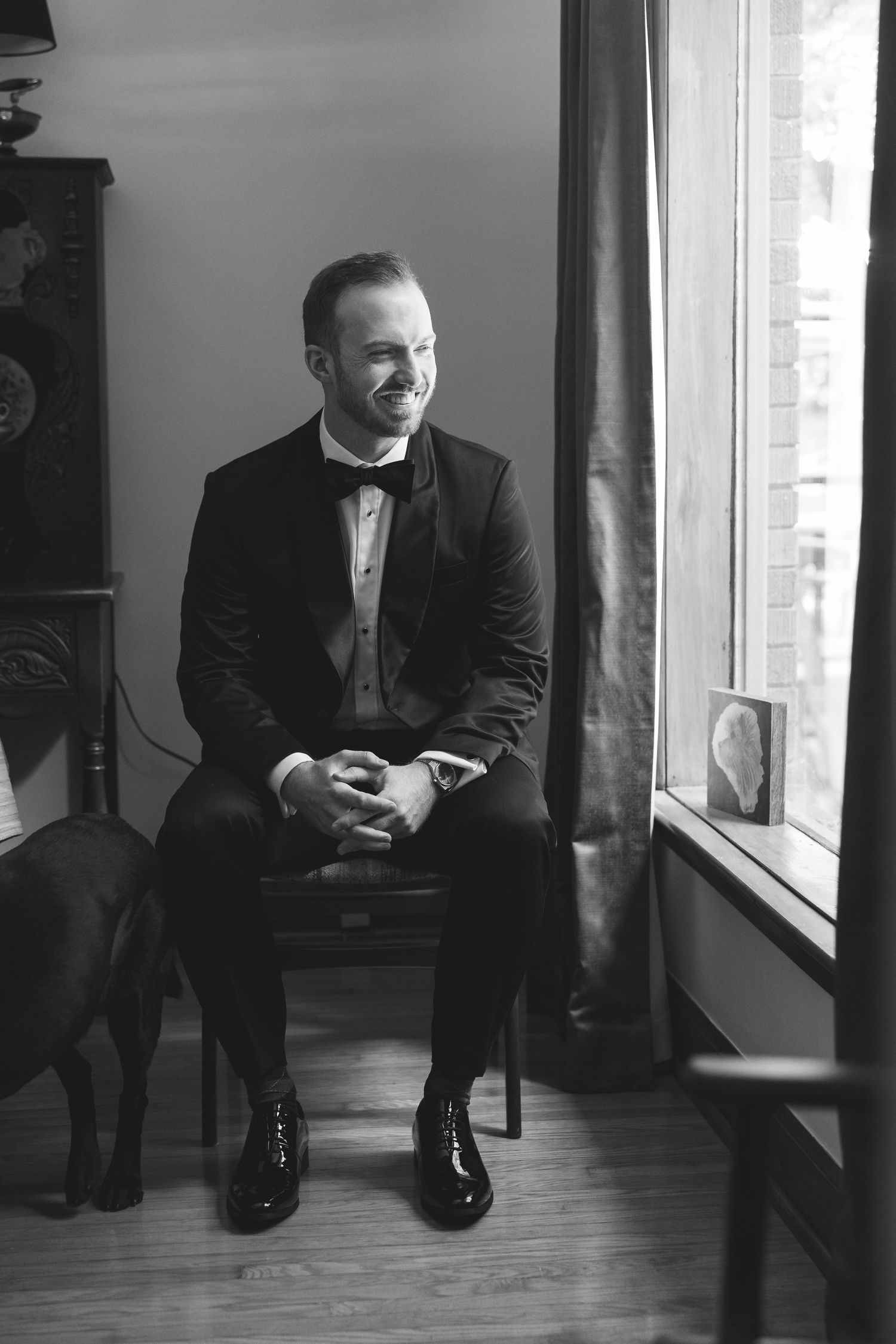 Black and white photo of a smiling groom in a tuxedo sitting by a window with a dog beside him.