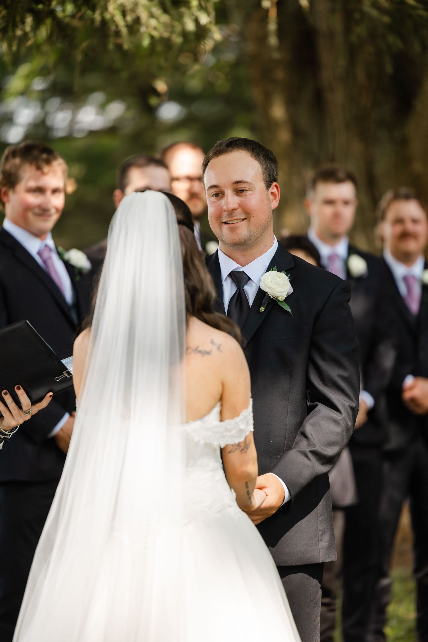 Groom smiles joyfully at bride while exchanging vows during outdoor wedding ceremony with groomsmen behind.