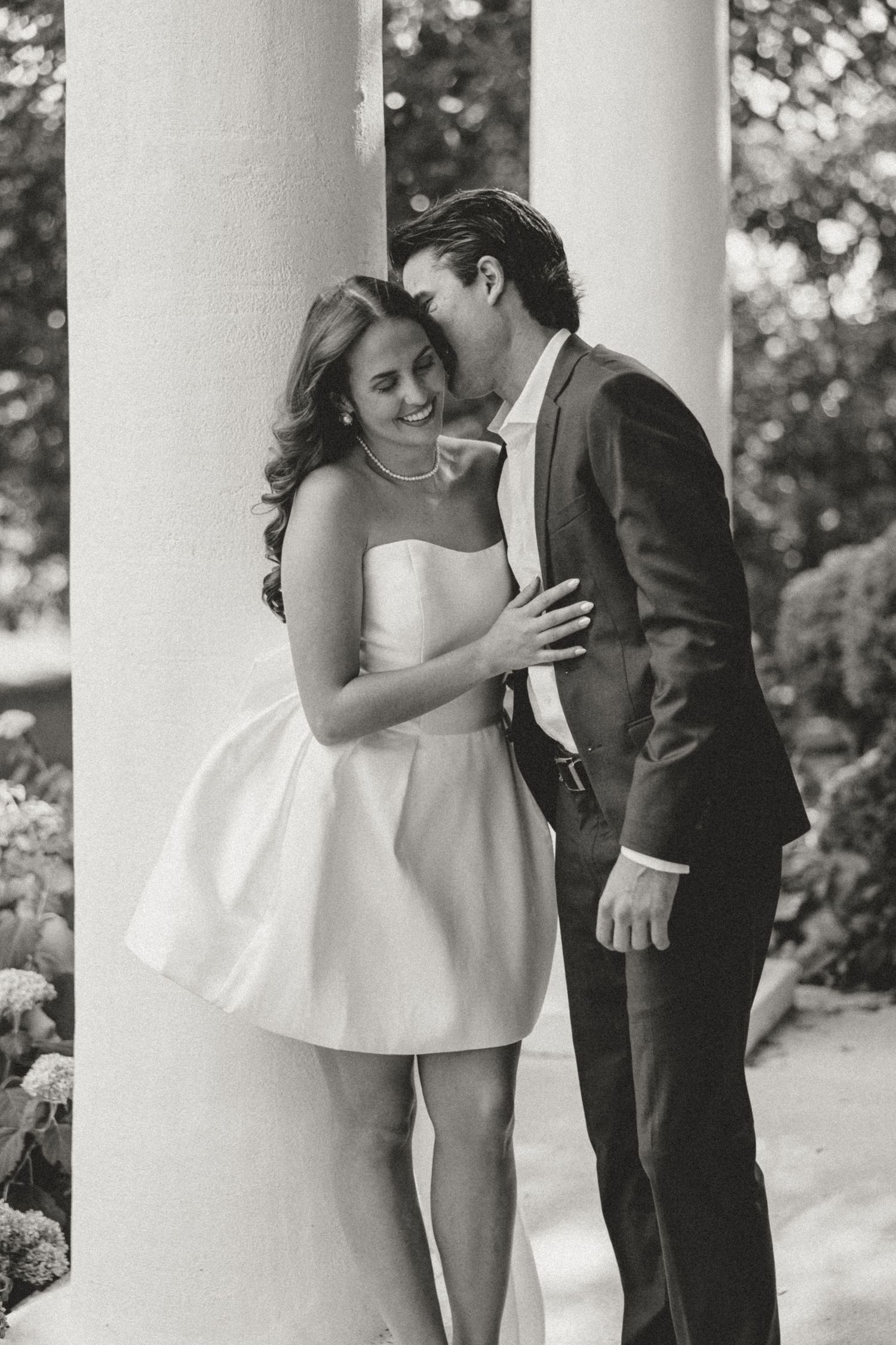 Black and white photo of a couple sharing a romantic moment between white columns, bride in short white dress.