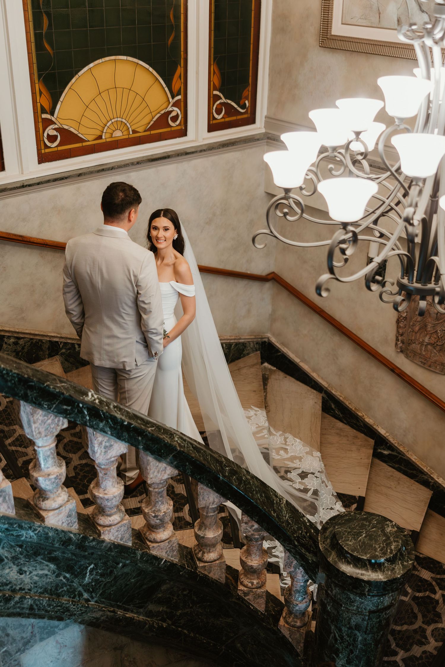 Bride in white gown and groom share romantic moment on grand marble staircase.