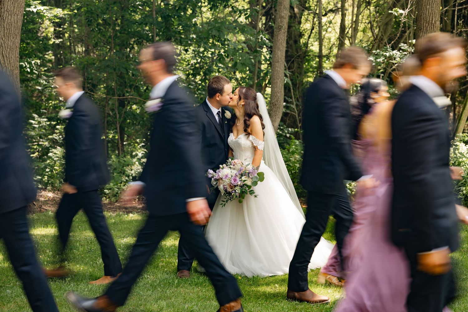 Bride and groom share a kiss while wedding party walks past in a lush garden setting.