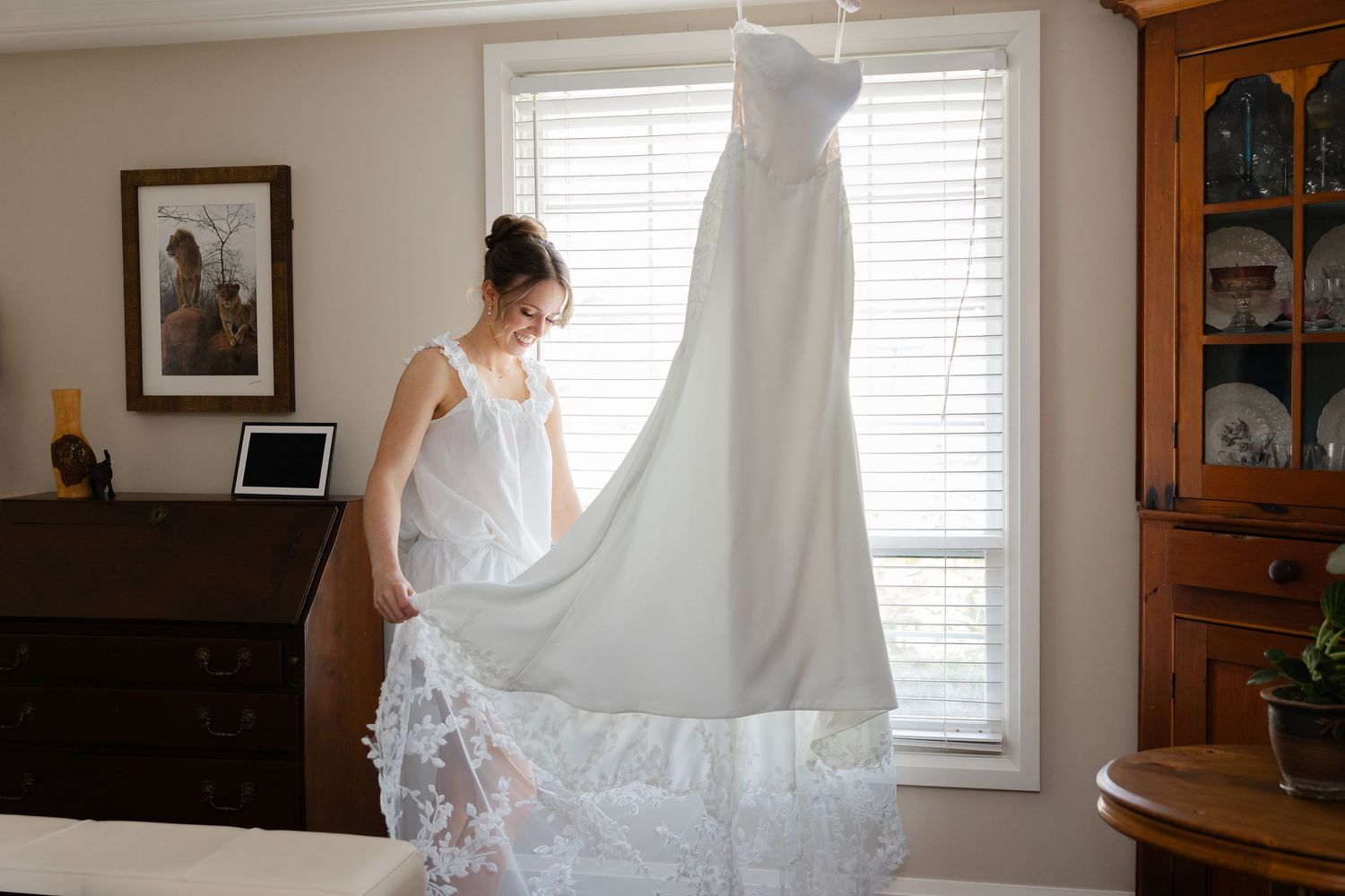 Bride admiring her elegant white wedding gown hanging by a window in a warmly decorated room.