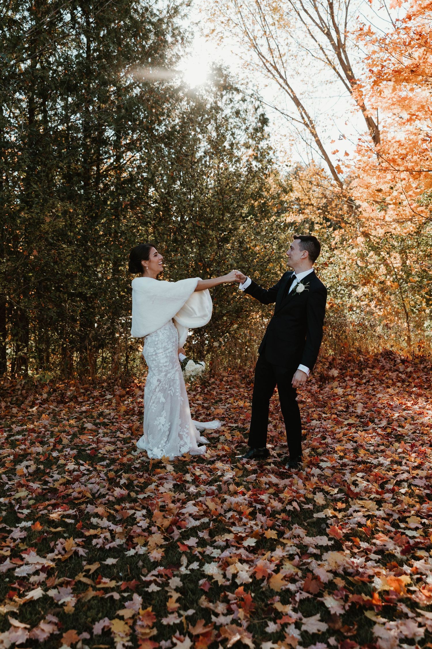 Bride and groom dance among vibrant autumn leaves in a sunlit forest wedding portrait.