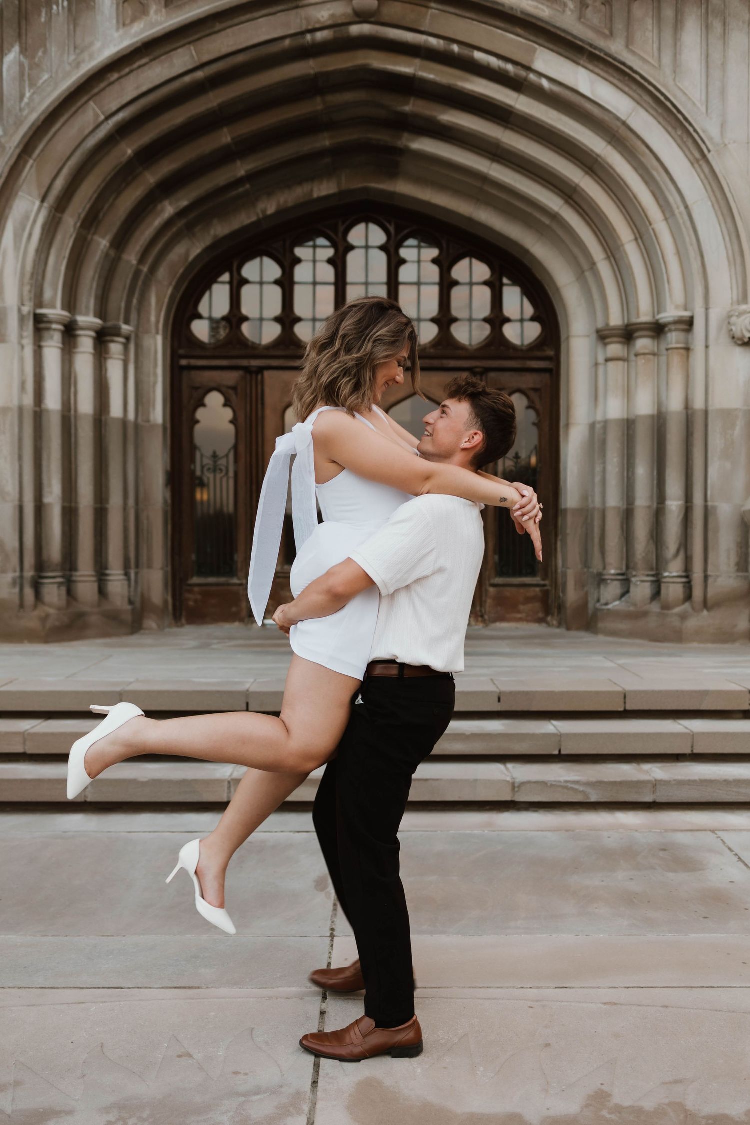 Man lifting woman in white dress in front of a grand Gothic stone archway entrance.