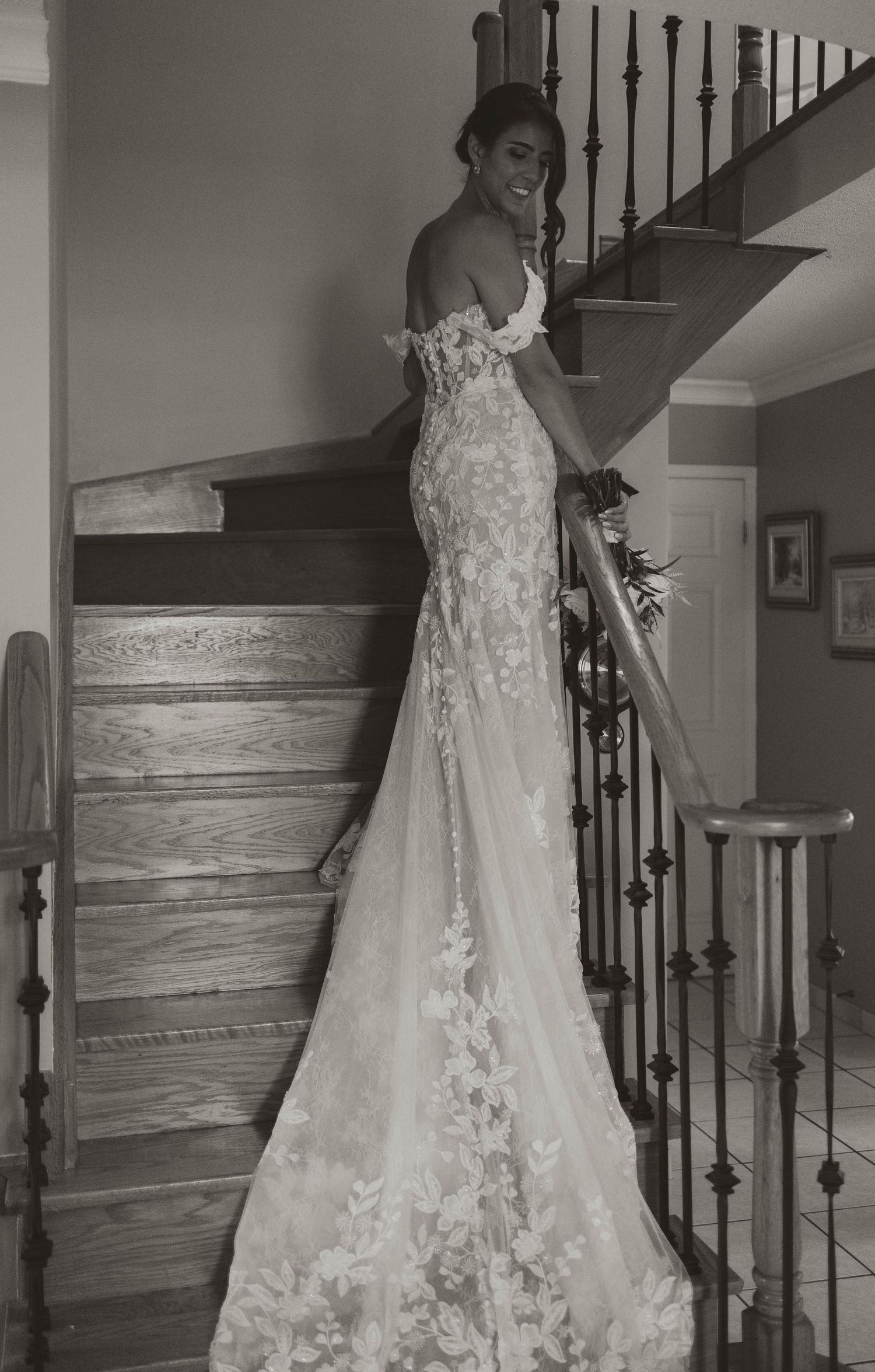 Elegant bride in floral lace off-shoulder gown poses on staircase in stunning black and white photo.