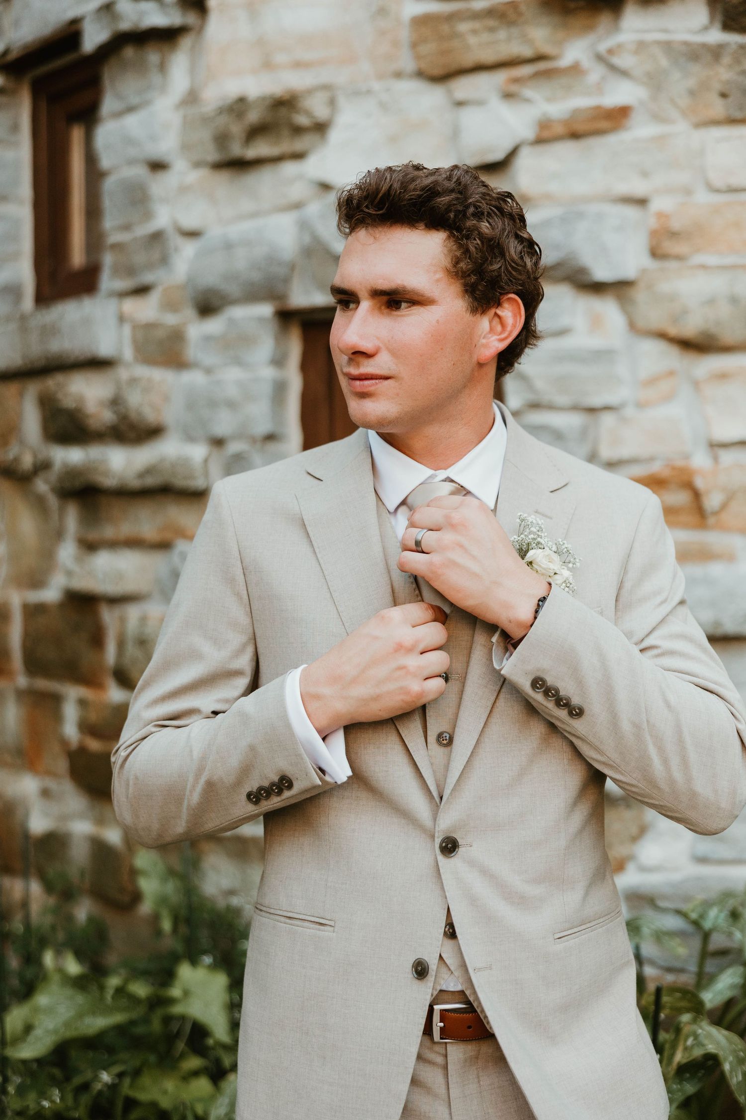 Groom in beige suit adjusting tie outside rustic stone wall venue.