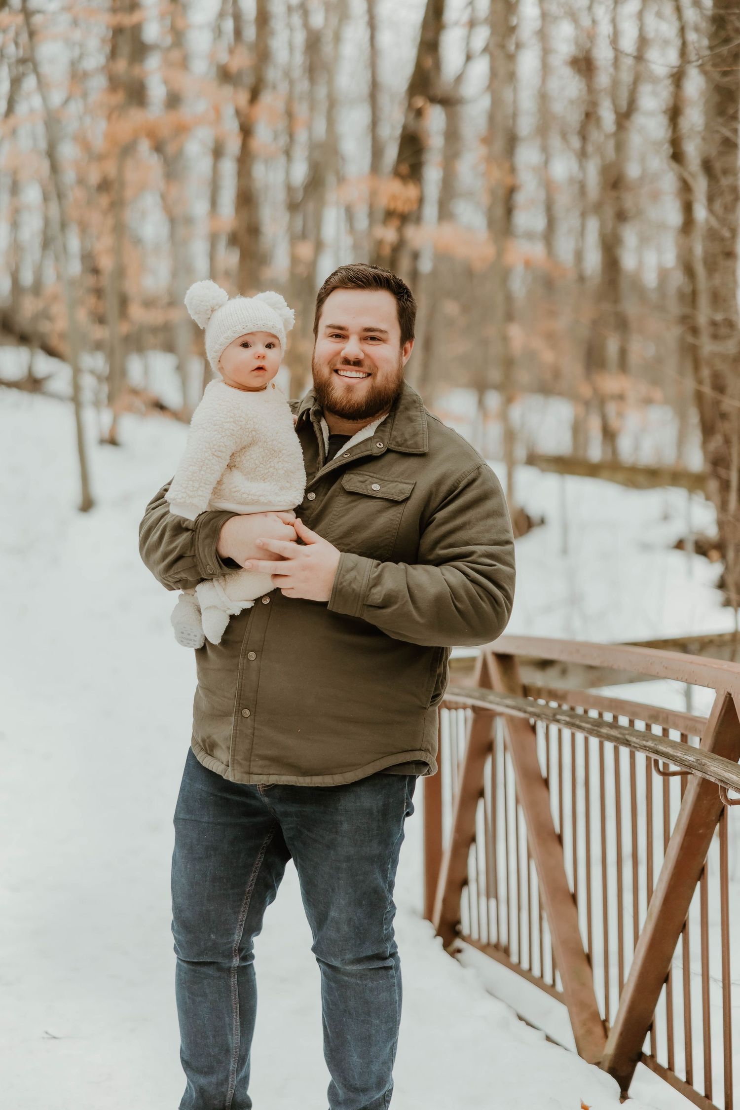 Smiling bearded man in green jacket holding baby in white knit hat on snowy bridge.