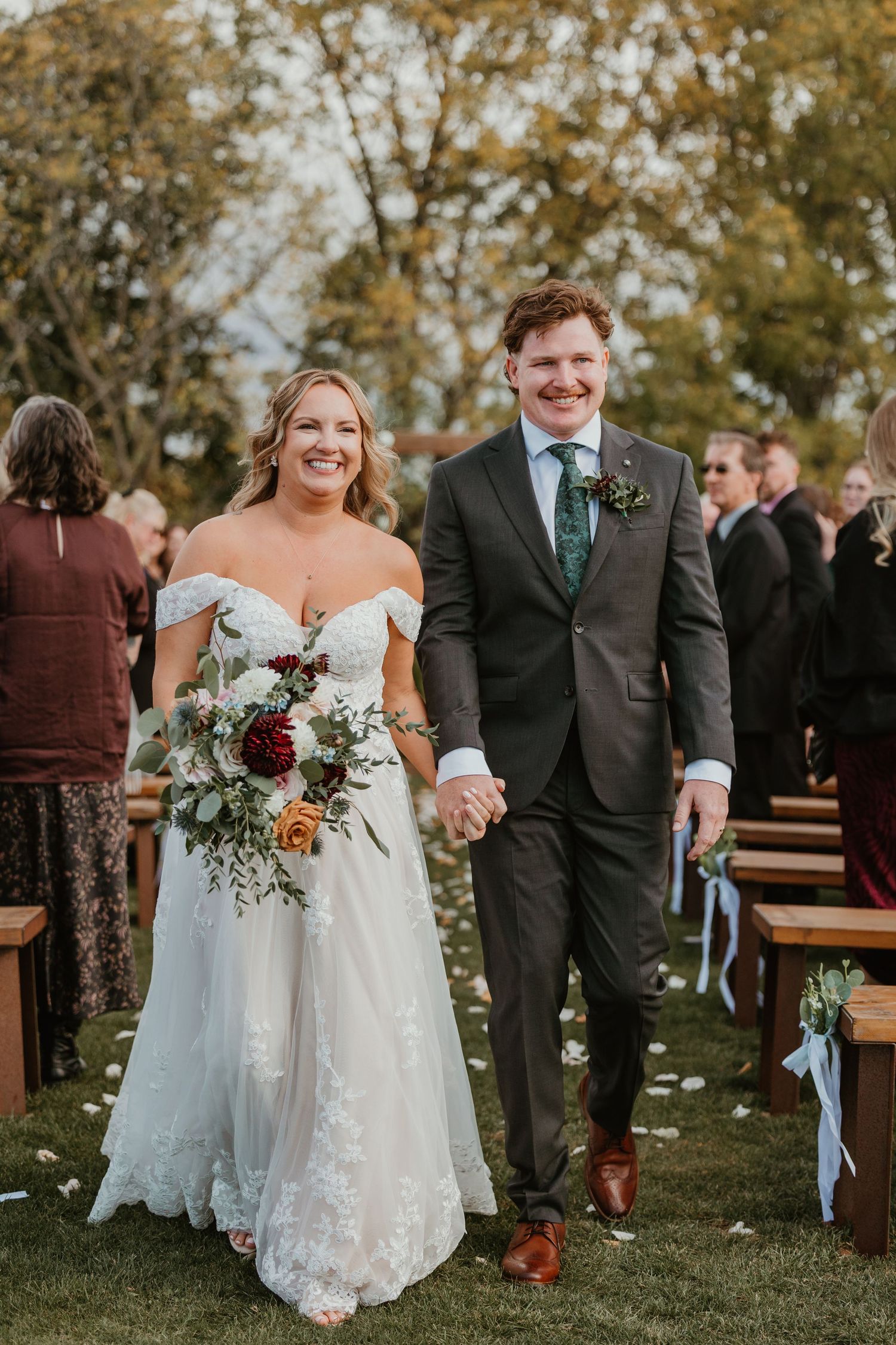 Smiling newlyweds walking down outdoor wedding aisle, bride in lace off-shoulder gown holding colorful bouquet.