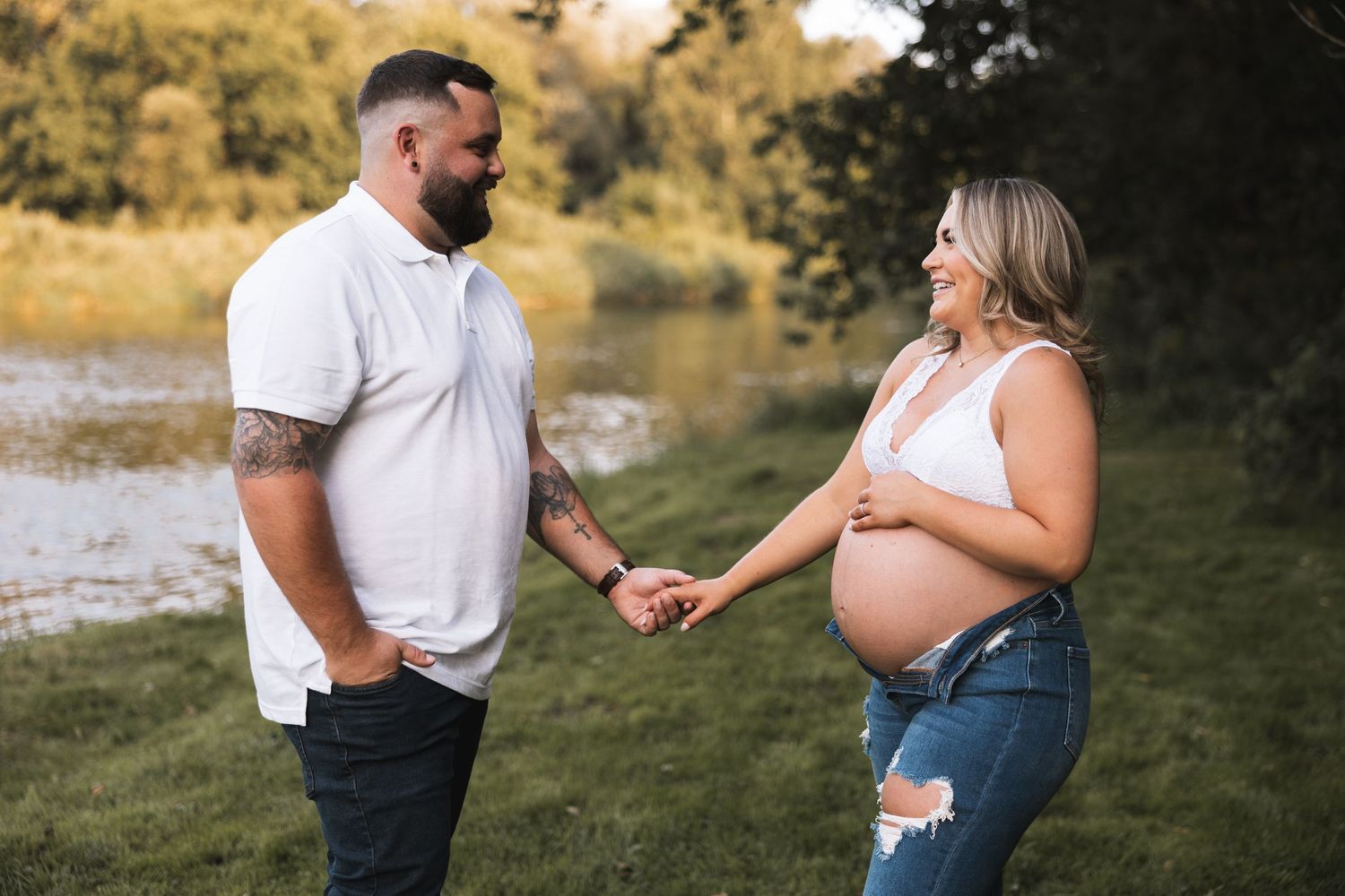 Expecting couple holding hands and smiling outdoors near a river.