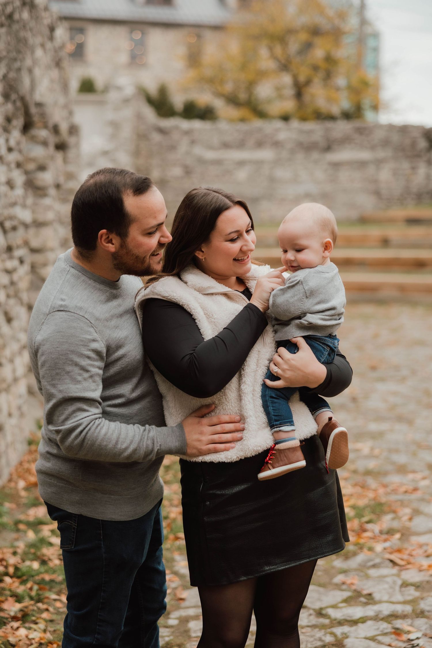 Happy family of three with parents holding baby boy, smiling together near a stone wall in autumn.