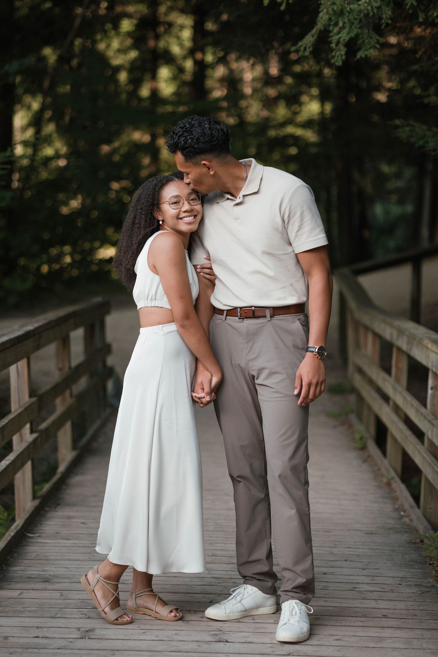 Couple holding hands on a wooden bridge in a forest, woman in white dress smiling at camera.