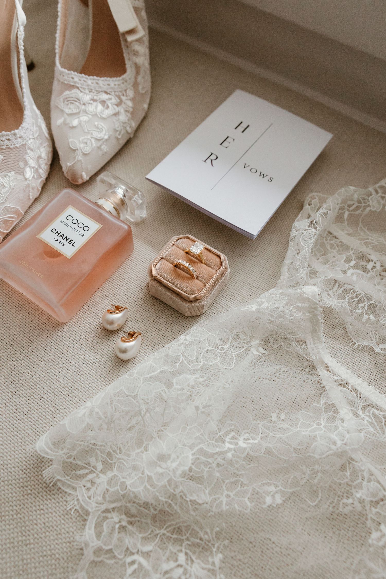 Bridal flat lay featuring Chanel perfume, gold rings, pearl earrings, lace veil, and Her Vows booklet.