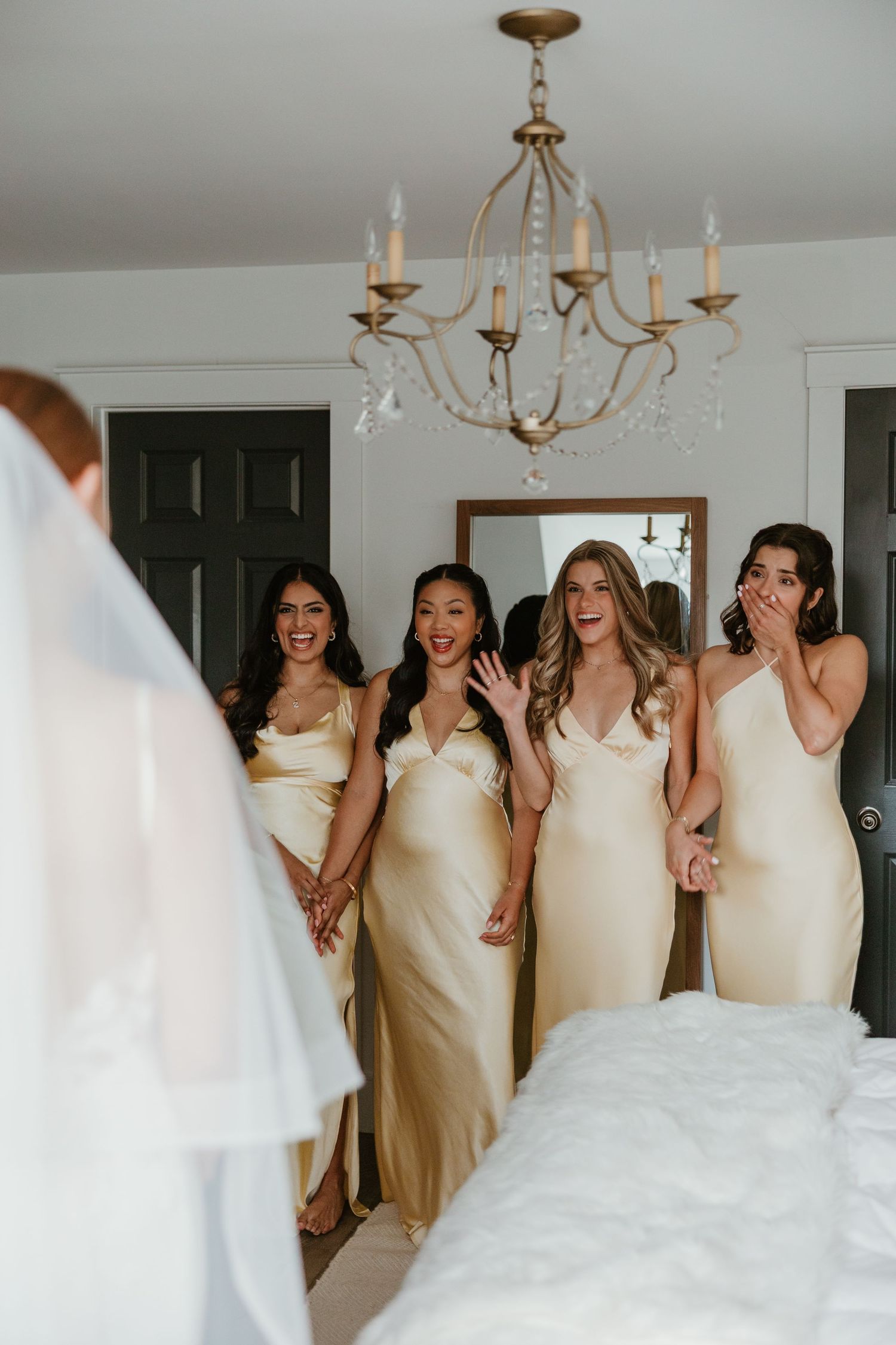 Four bridesmaids in gold satin dresses react with excitement seeing bride in her wedding gown.
