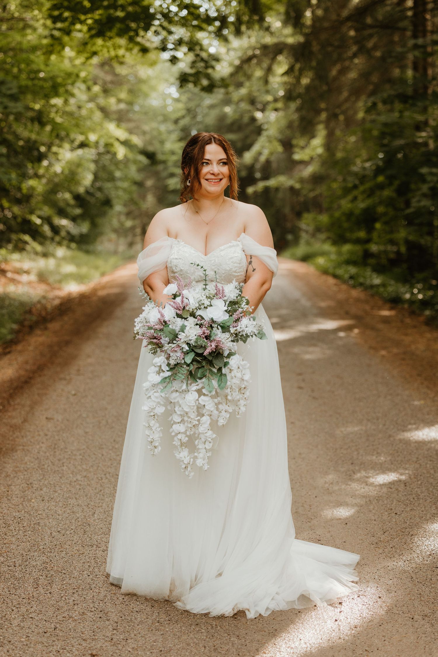 Bride in off-shoulder white gown holding cascading floral bouquet on a tree-lined dirt path.