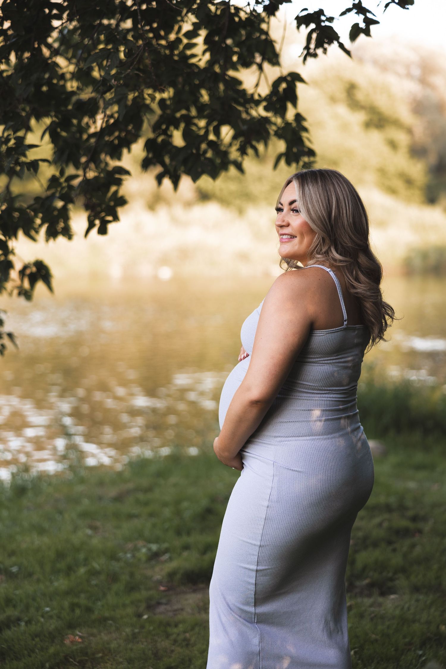 Smiling pregnant woman in a grey dress poses by a serene lake during a maternity photoshoot.