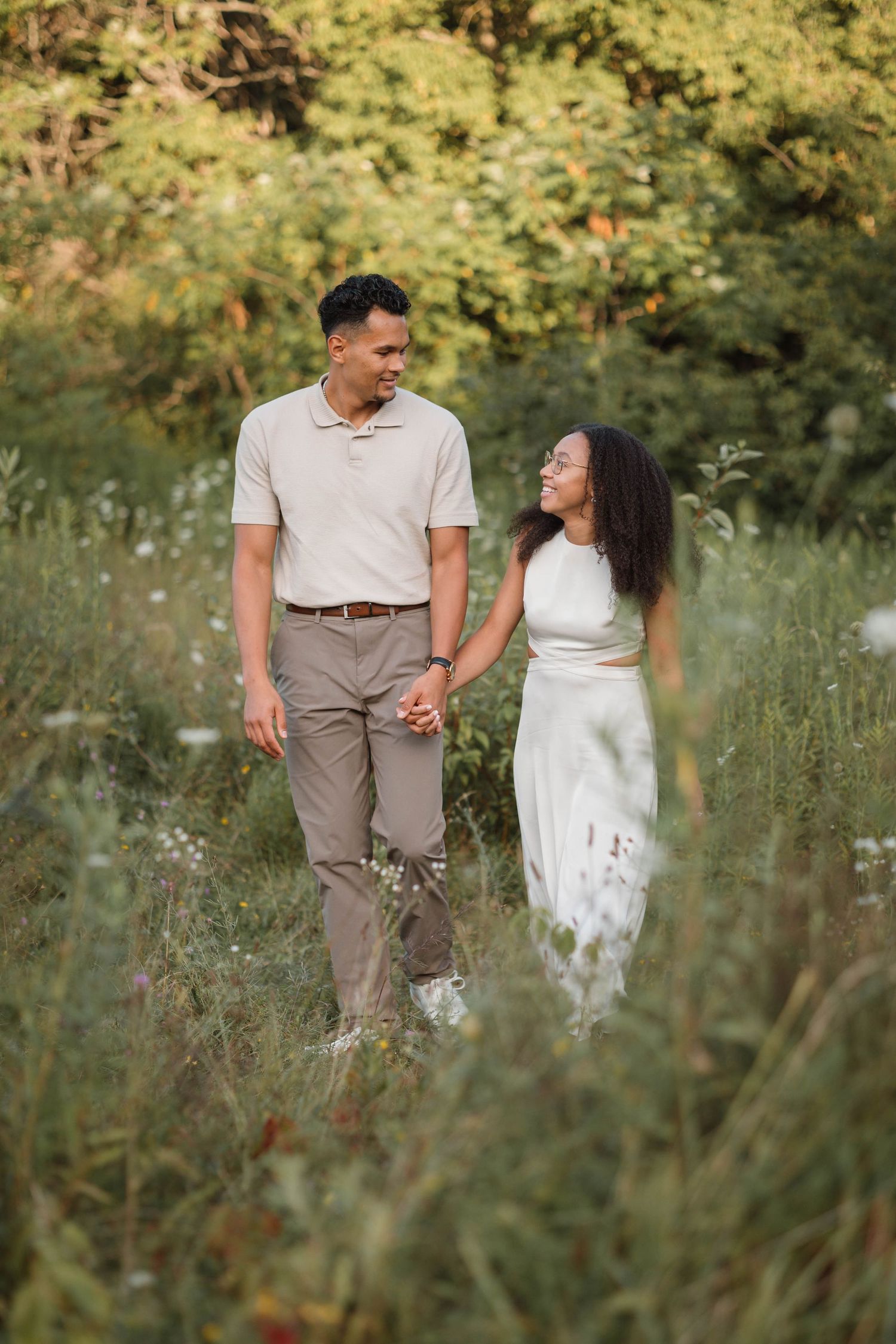 Couple walking hand in hand through a wildflower meadow, smiling at each other in golden light.