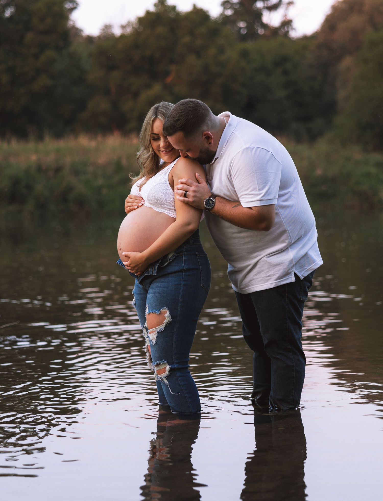 Couple standing in water, man kissing pregnant woman's head at sunset.