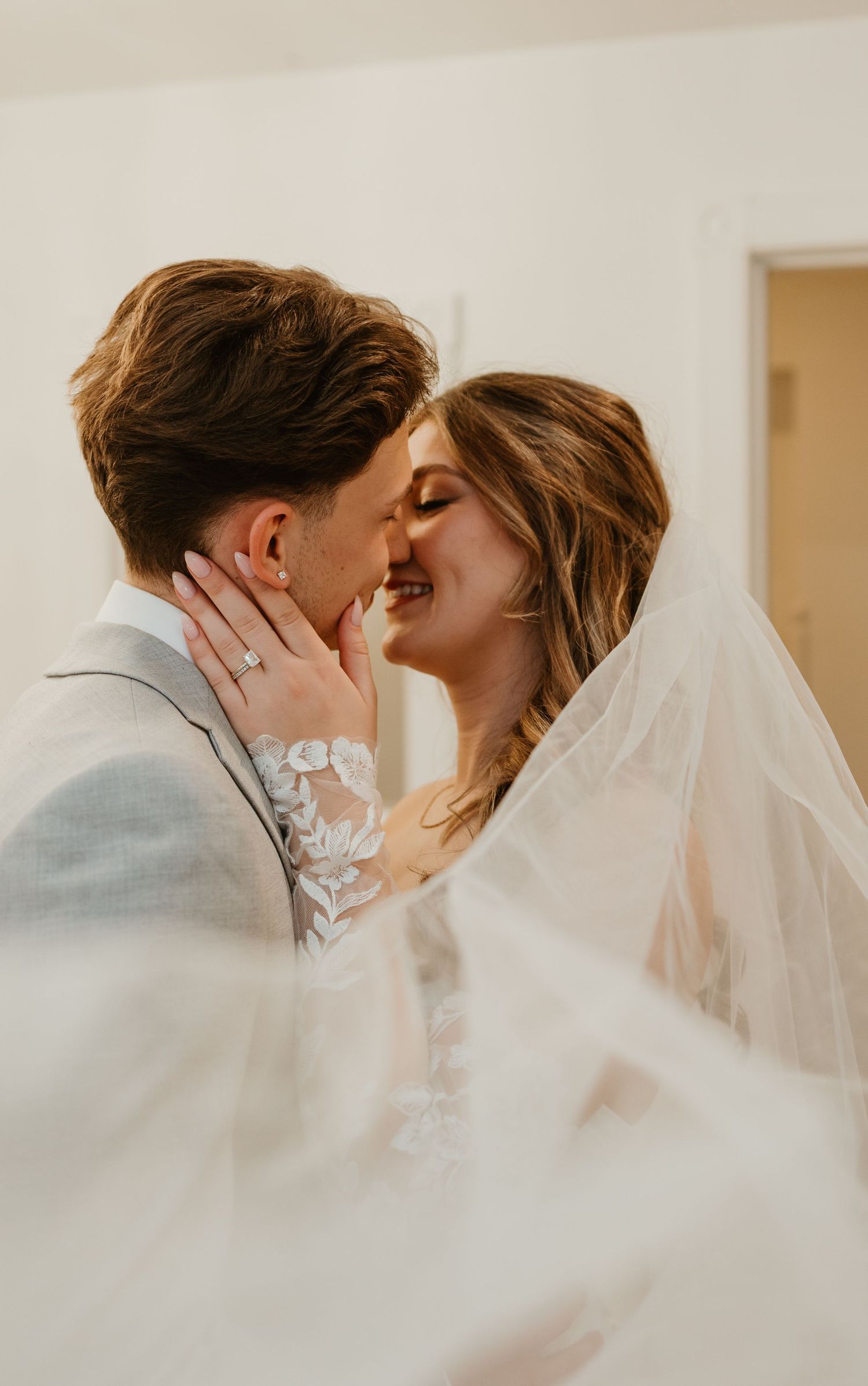 Bride smiling as groom leans in for a kiss, wrapped together under a flowing lace wedding veil.