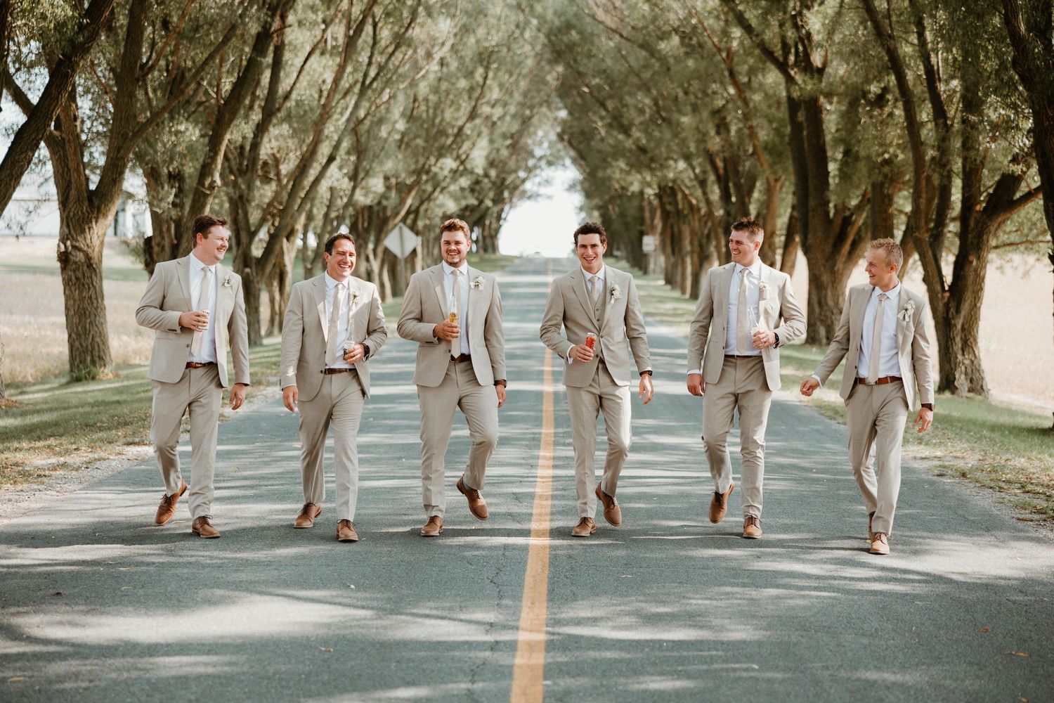 Six groomsmen in matching tan suits walk confidently down tree-lined road in outdoor wedding portrait.
