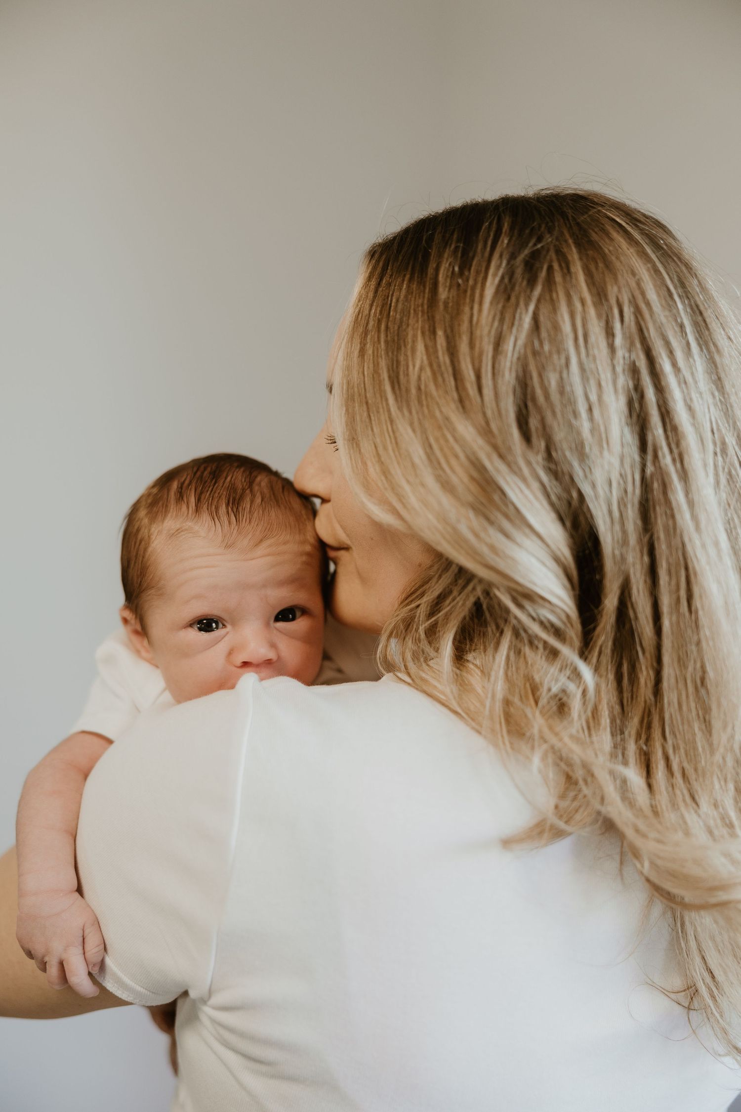 Blonde mother kisses her newborn baby on the cheek in a tender moment.
