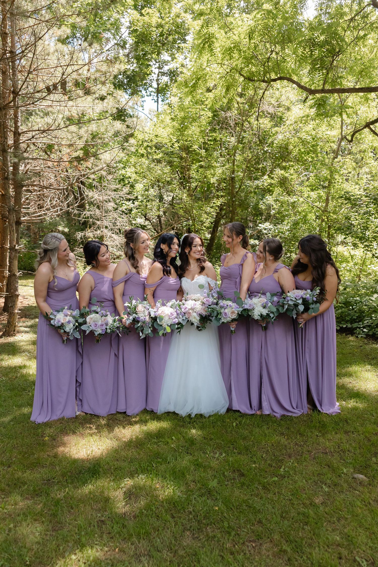 Bride with six bridesmaids in lavender gowns holding floral bouquets in a sunlit garden setting.