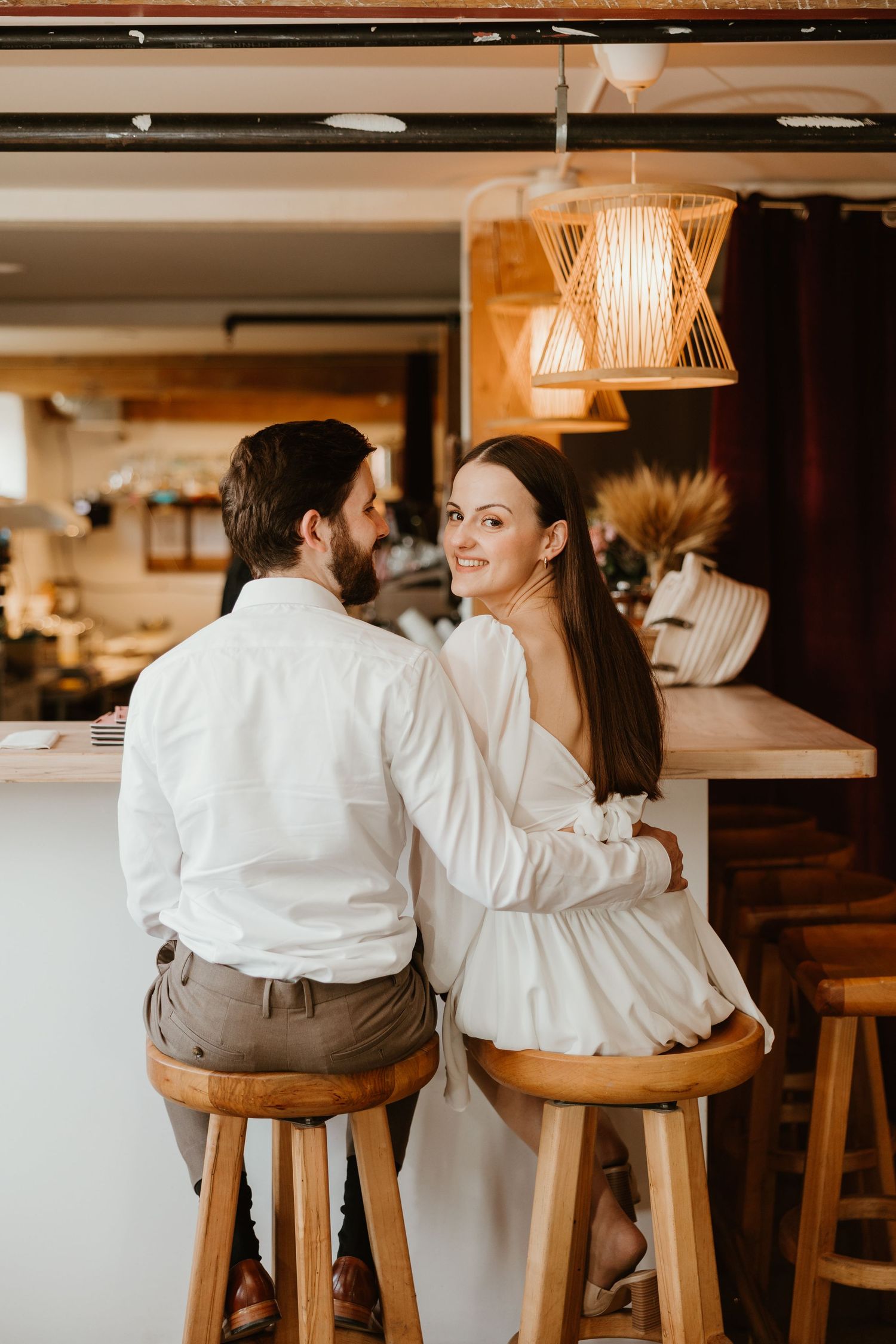 Smiling bride sitting at a bar counter with groom in a cozy restaurant engagement session.