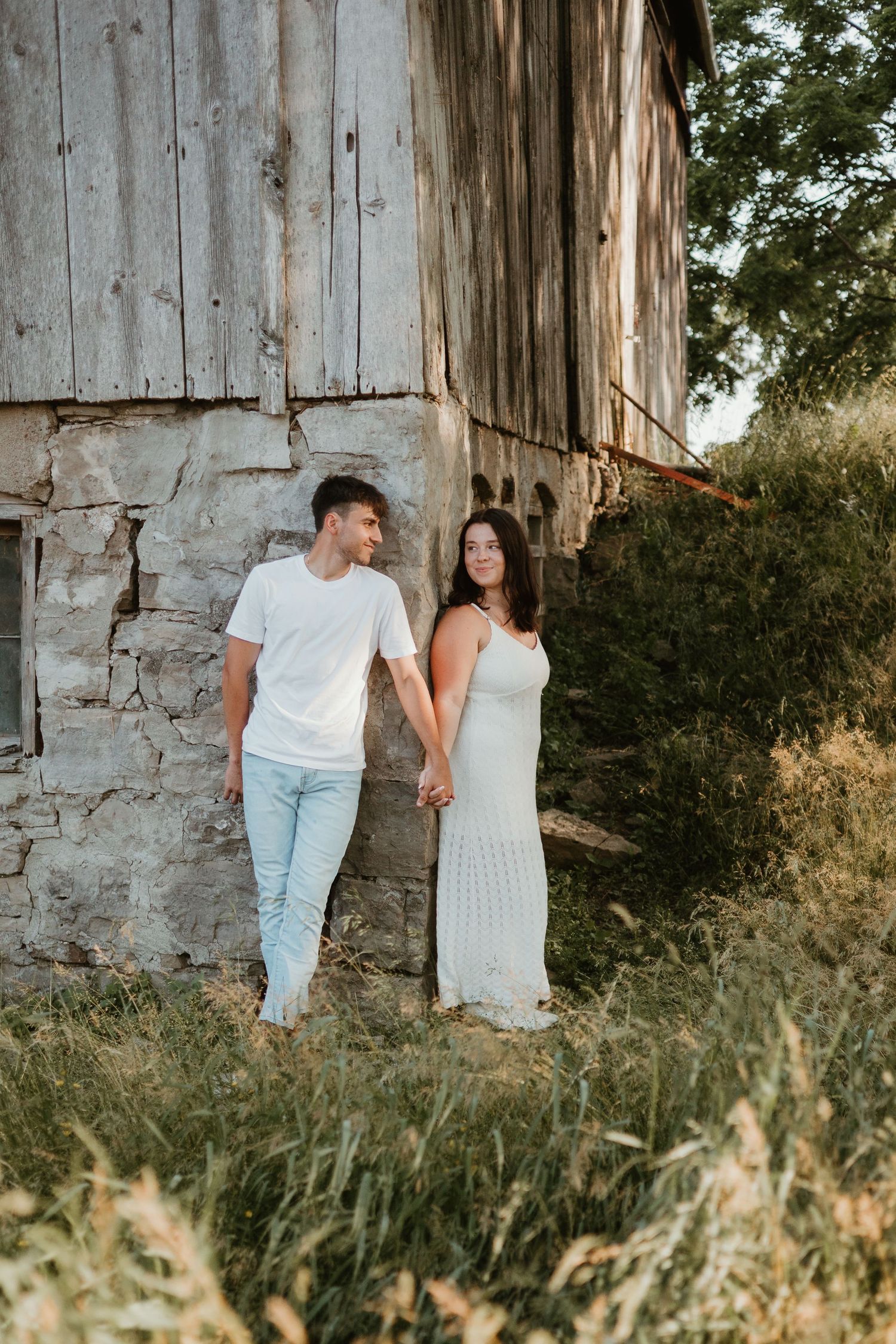 Couple holding hands and smiling at each other while leaning against a weathered stone barn.