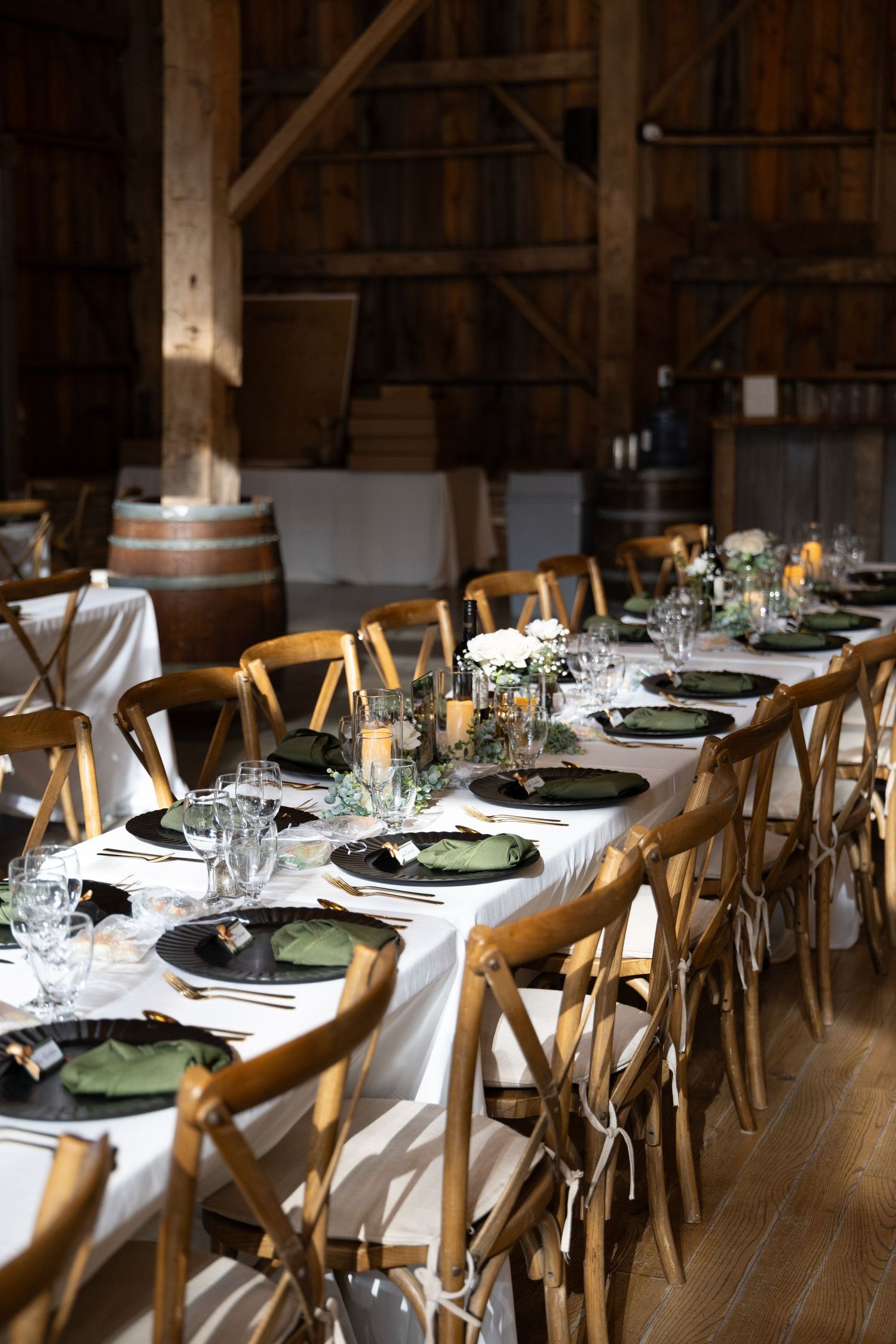 Long rustic wedding reception table with white linens, black plates, green napkins, candles and wooden chairs inside a barn venue.
