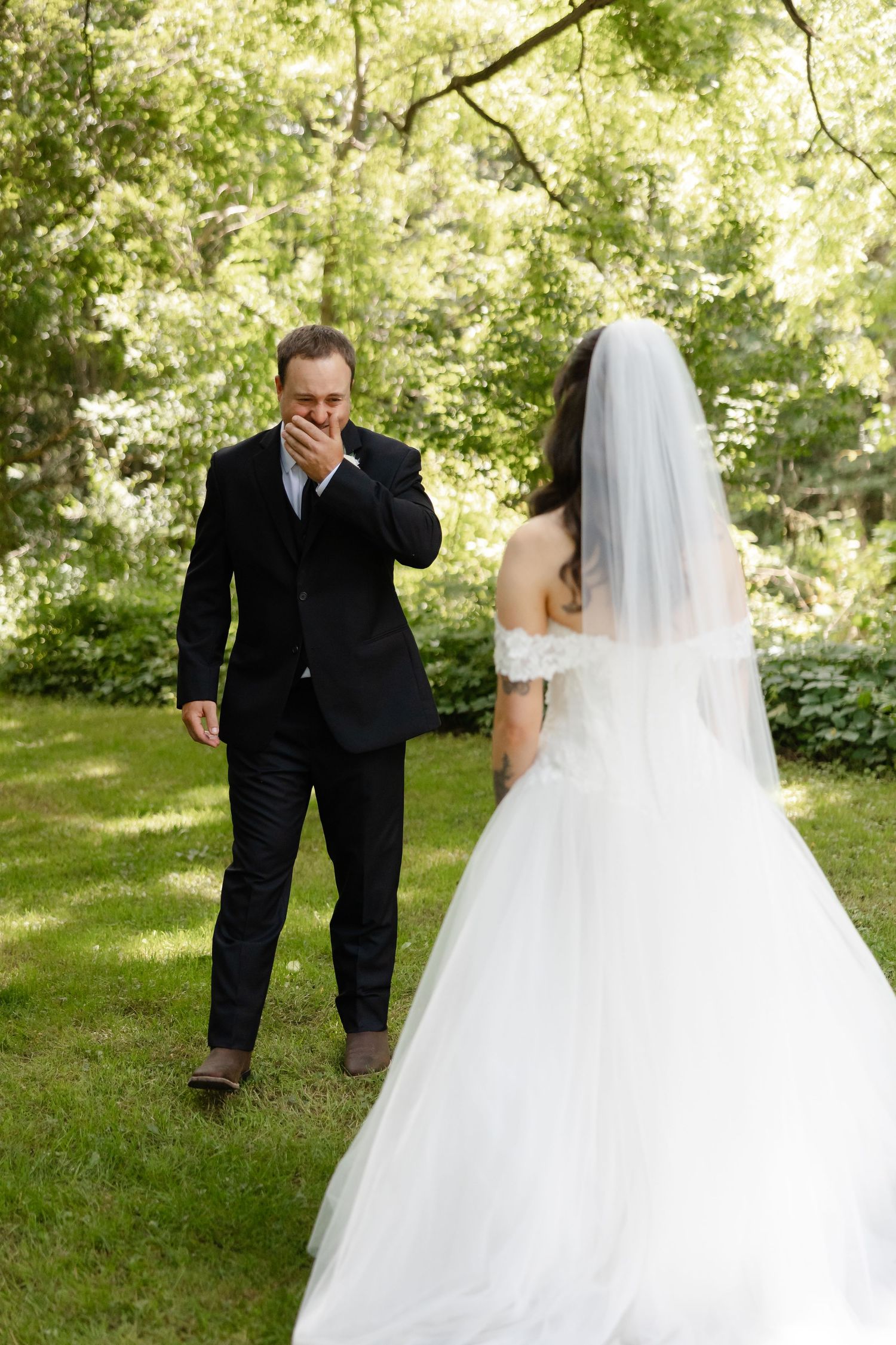 Emotional groom covers his mouth in awe seeing bride in white wedding gown for first look in lush garden.