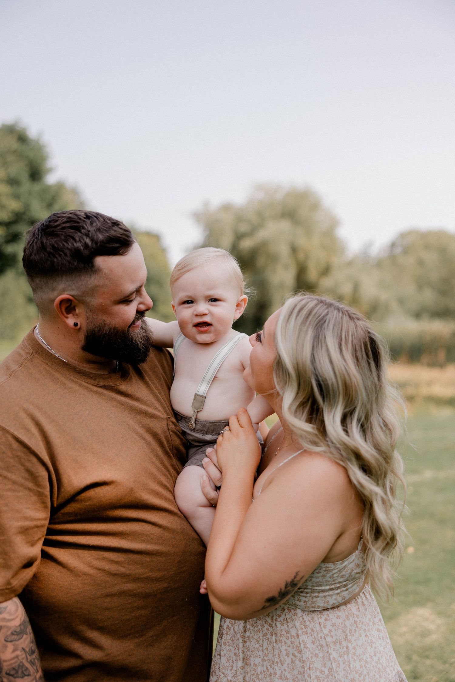 Couple outdoors in park smiling at happy baby in suspenders on sunny day.