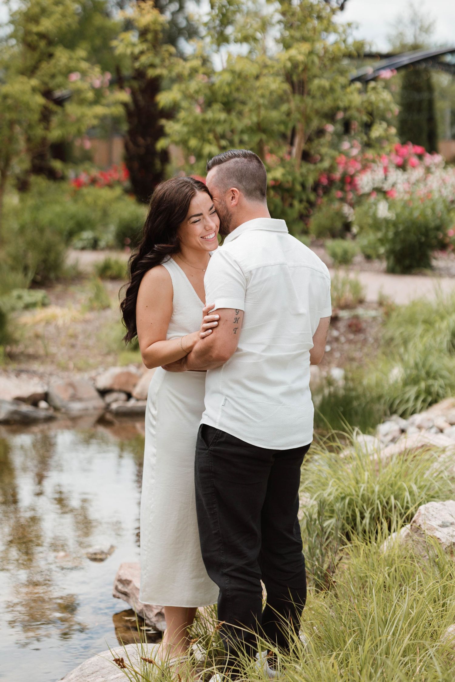 Couple embracing and smiling beside a garden pond surrounded by colorful flowering plants.