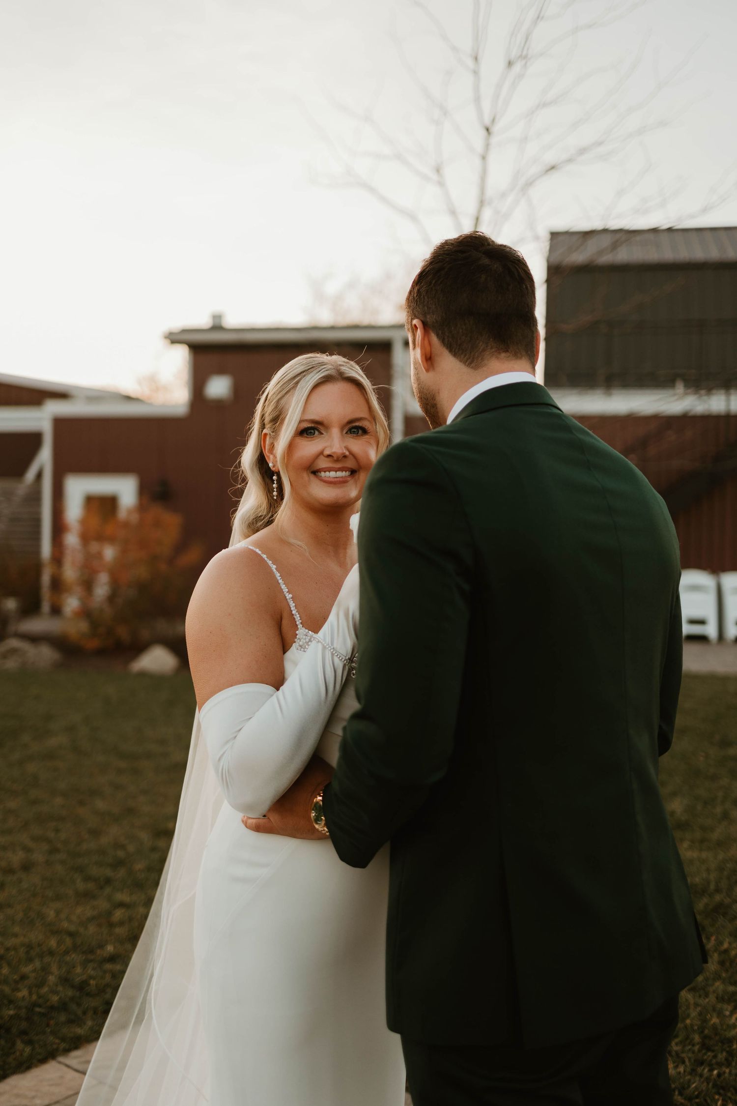 Smiling bride in white gown with wrap glances back at groom in dark suit outdoors at rustic wedding venue.