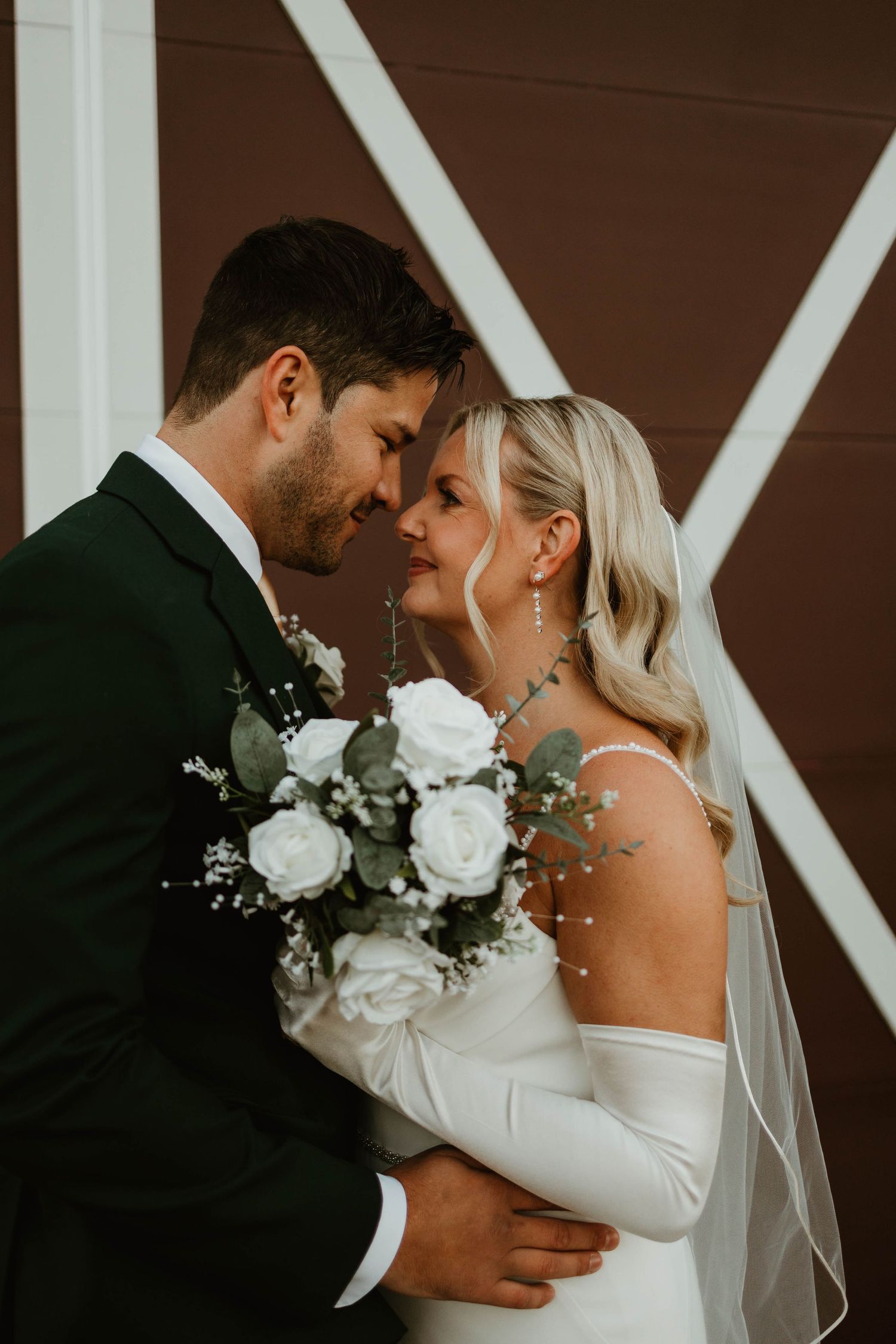 Bride and groom share an intimate moment, touching foreheads in front of a brown barn door.
