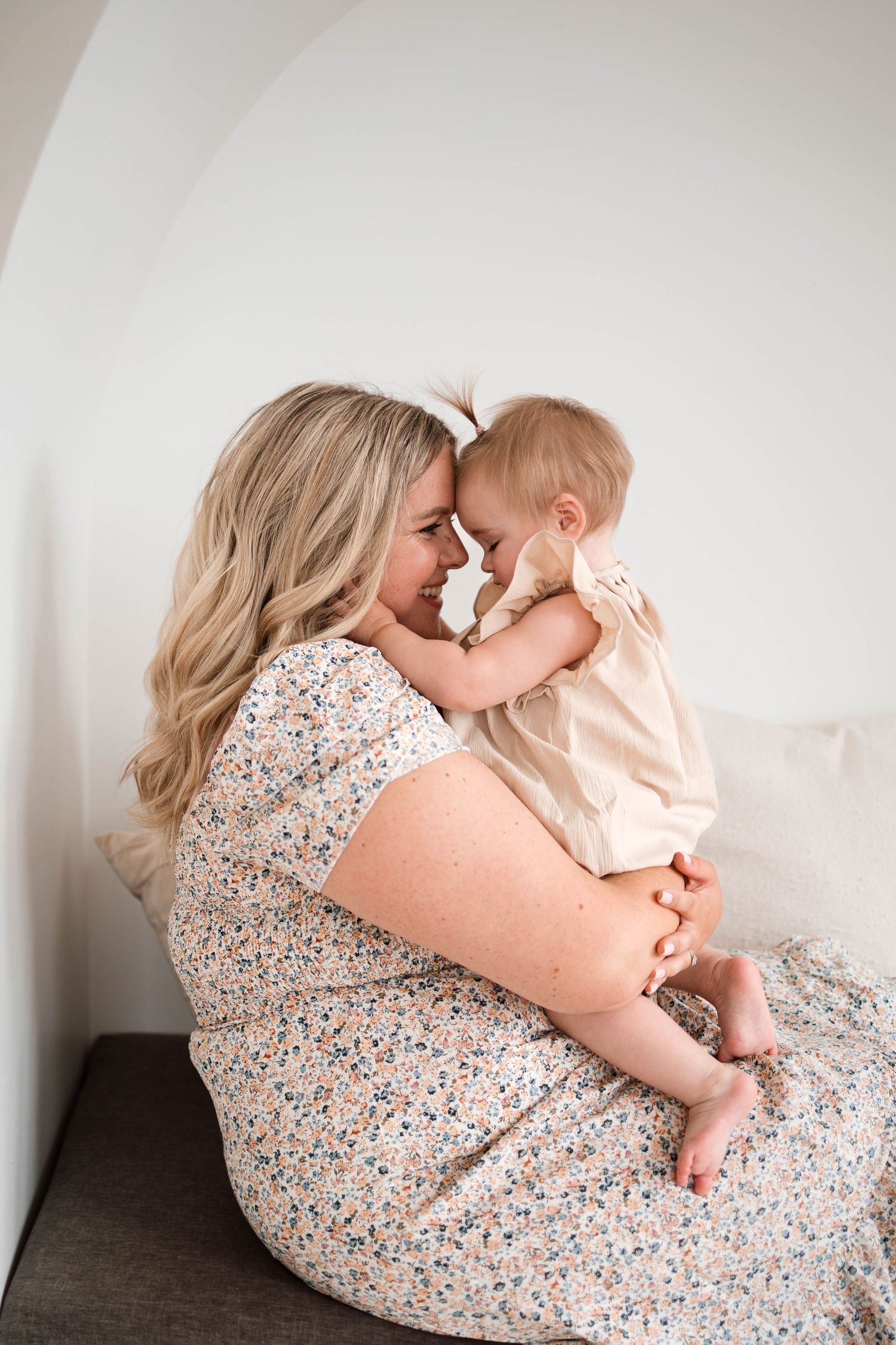 Blonde mother in floral dress sitting and touching foreheads with her baby in a bright white room.