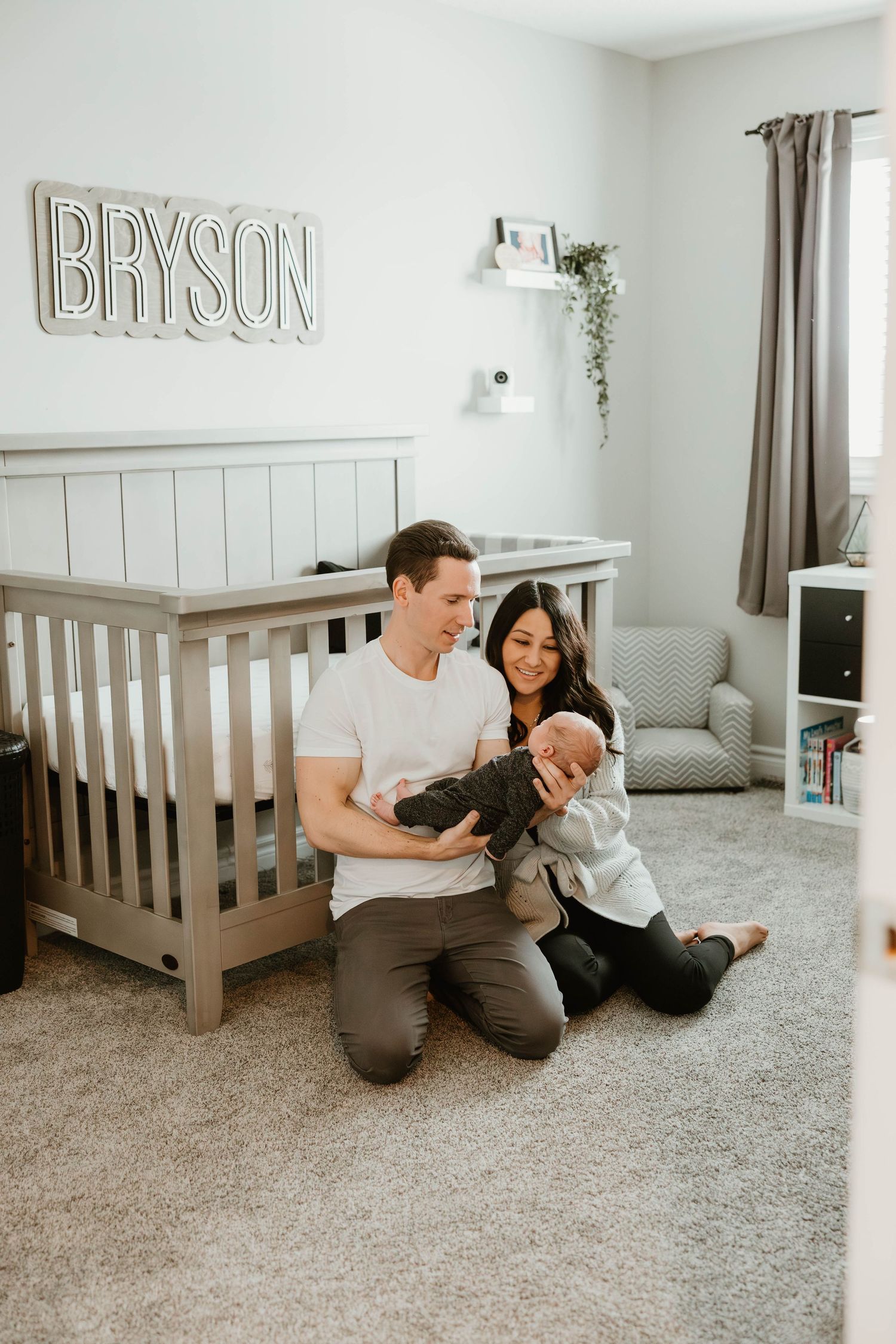 Happy parents sit on nursery floor holding newborn baby in front of crib with BRYSON name sign.