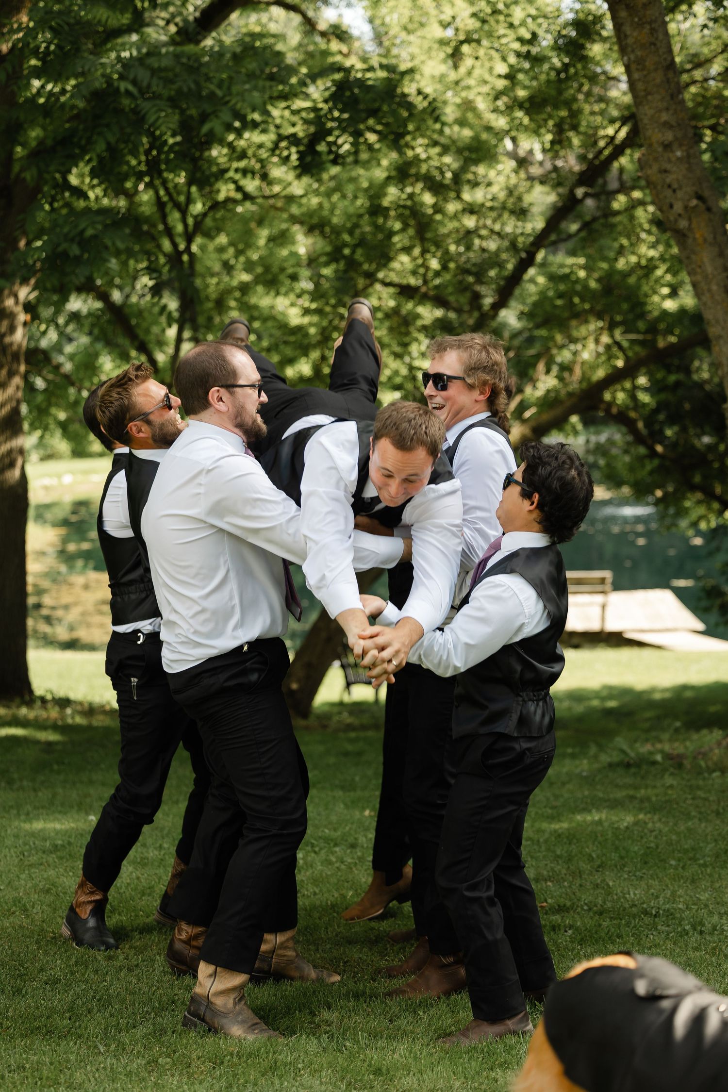 Groomsmen in black vests playfully lifting groom in celebration during outdoor wedding reception.