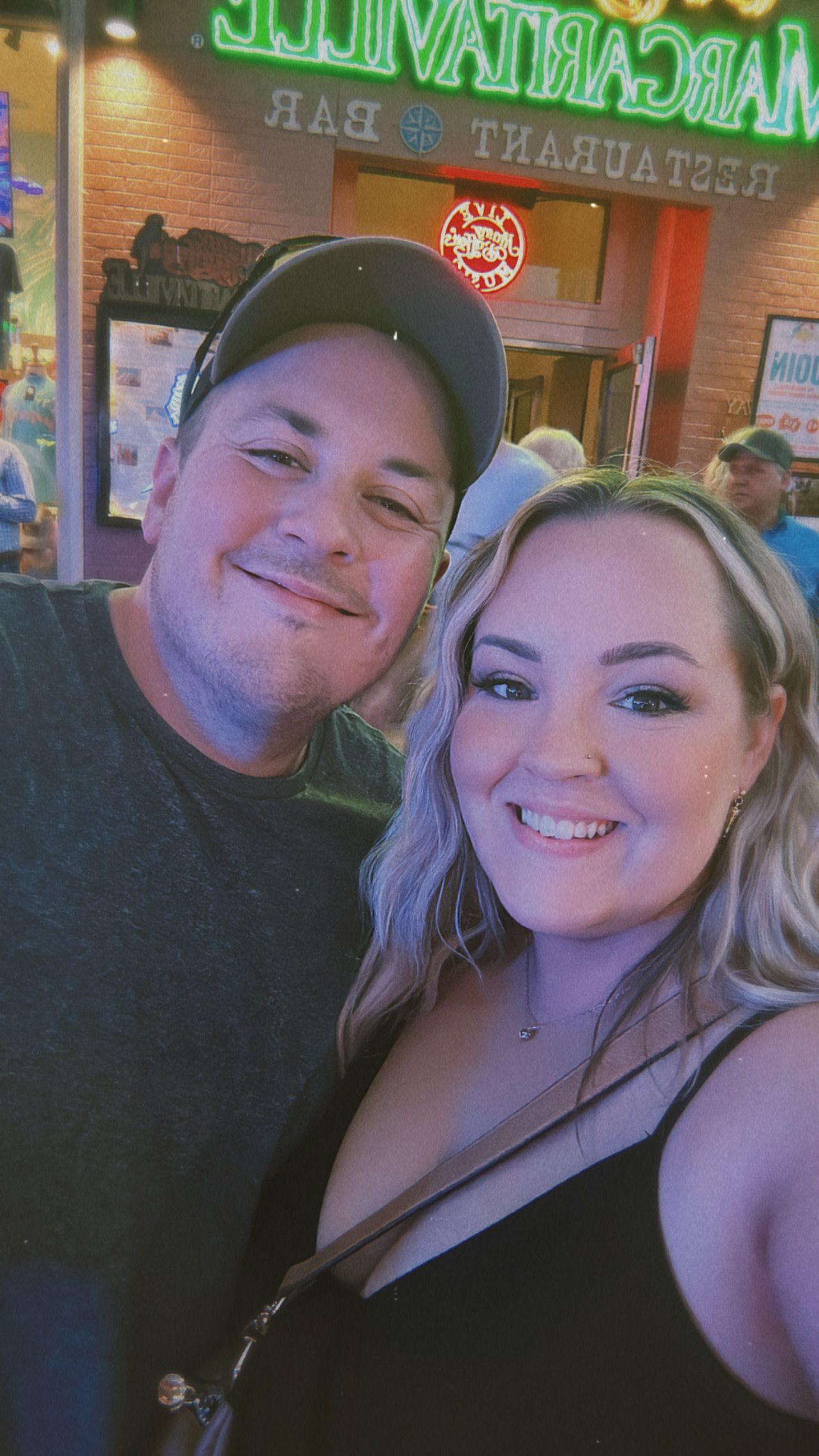 A couple smiles together outside a Margaritaville Restaurant and Bar at a lively entertainment venue.