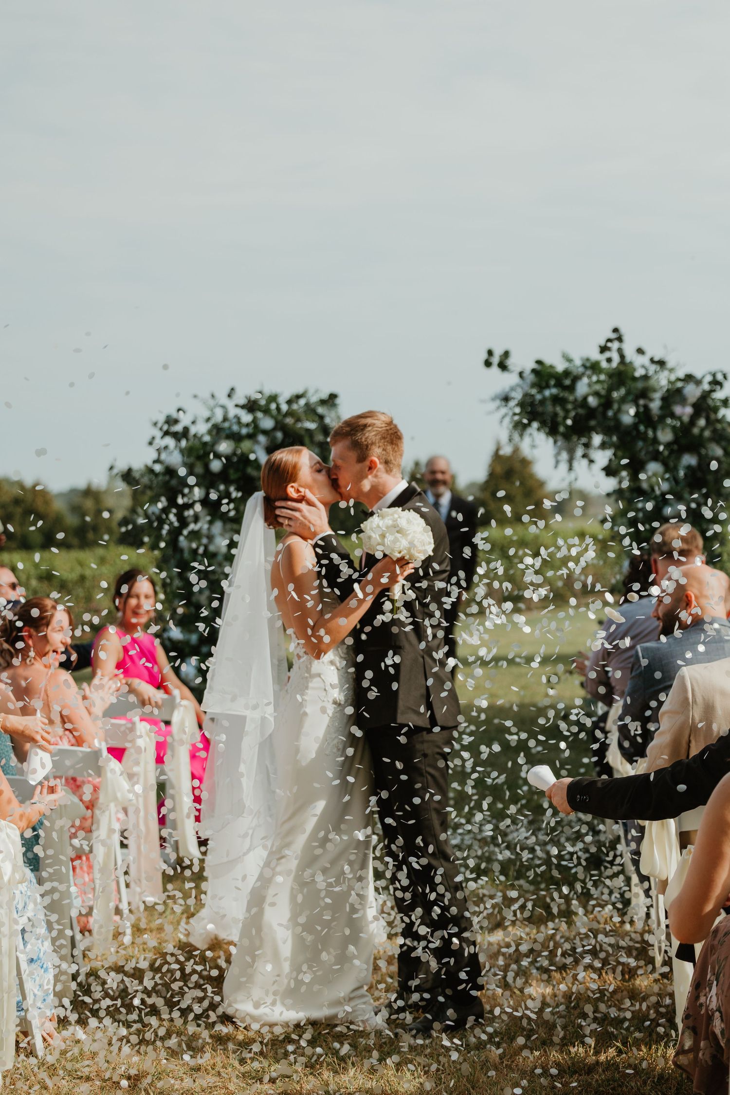 Newlyweds sharing first kiss as confetti fills the air during outdoor wedding ceremony celebration.