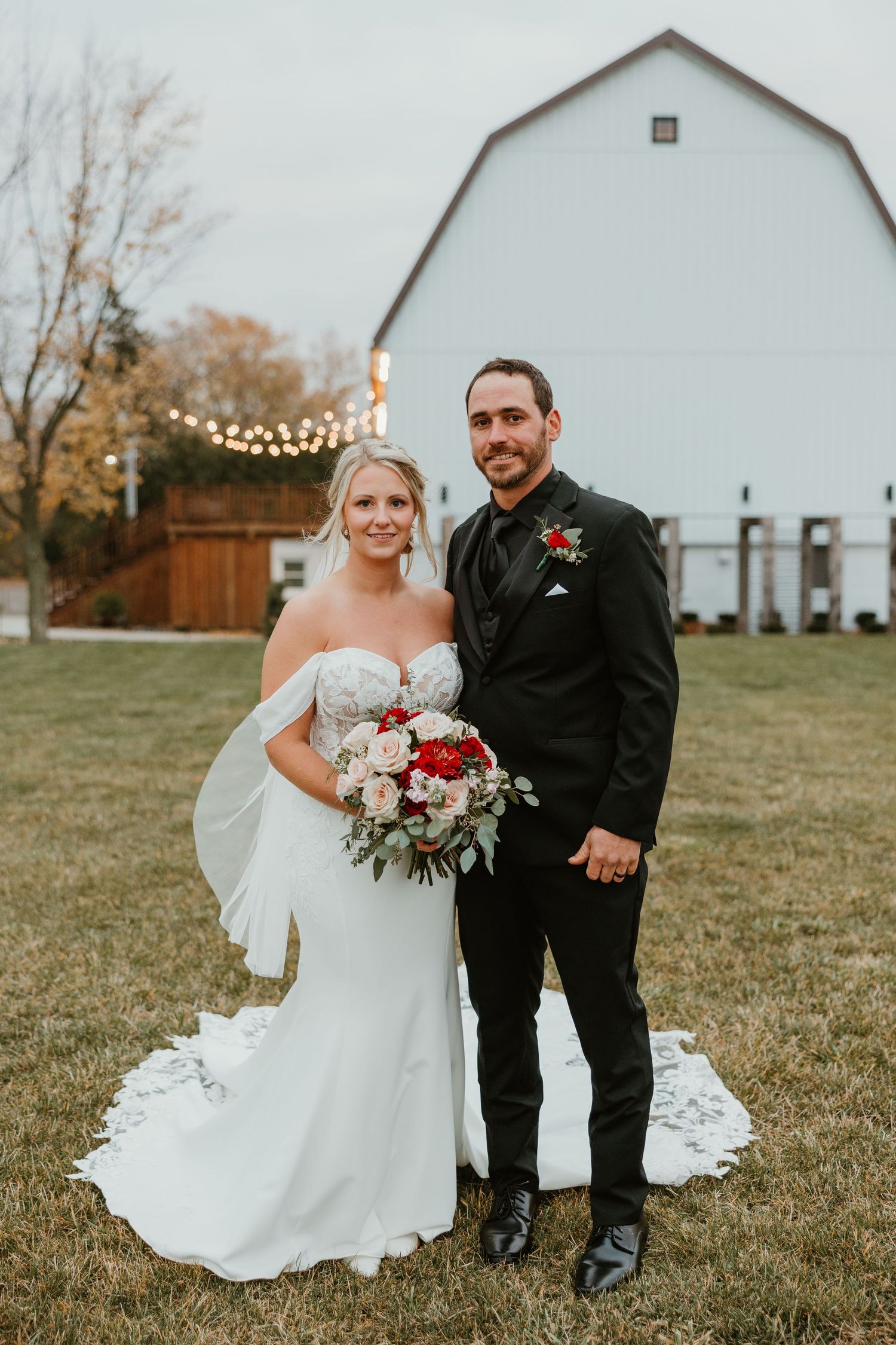 Bride and groom pose in front of a white barn with string lights at autumn wedding venue.